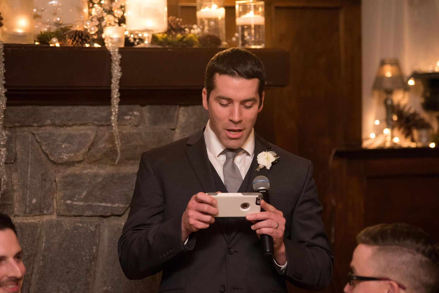 Man in suit reading speech at wedding reception near stone fireplace with candles.