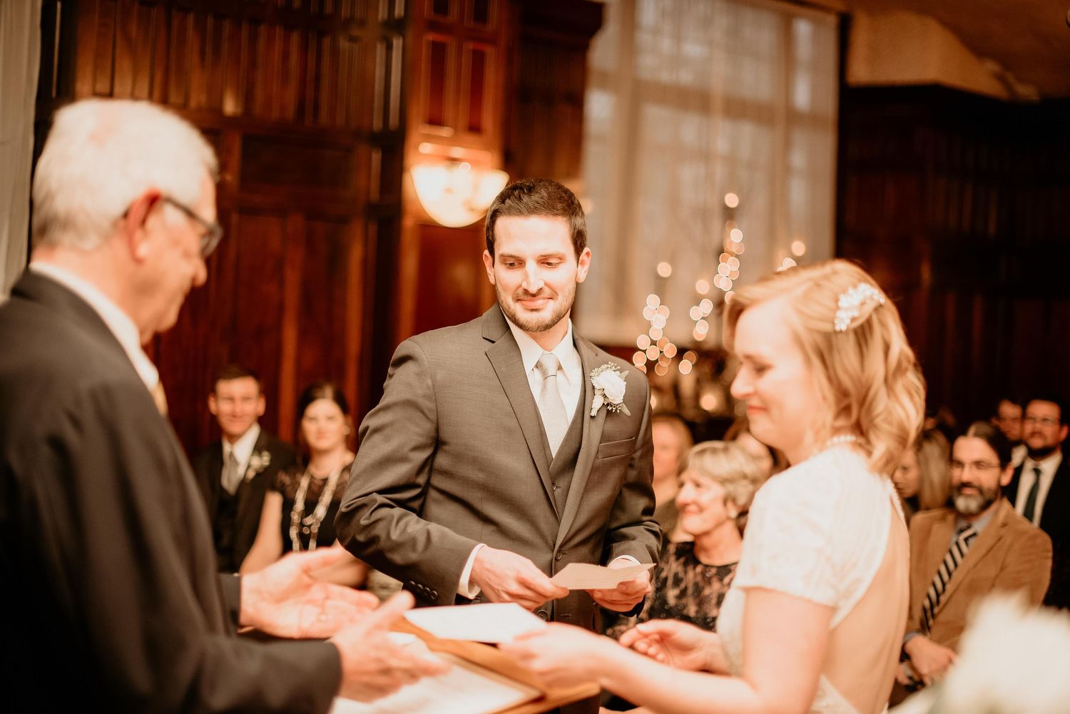 Smiling groom exchanges vows with bride during elegant indoor wedding ceremony with guests in background.