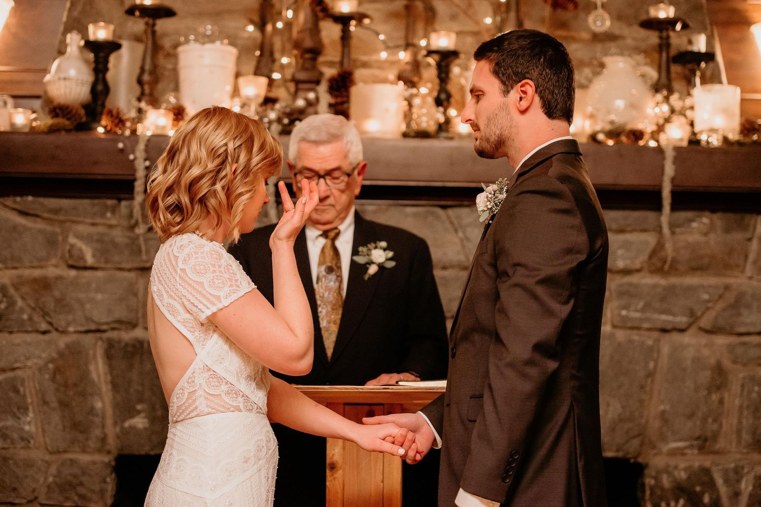 Emotional bride wiping tears during intimate candlelit wedding ceremony while groom holds hands with officiant at stone fireplace.