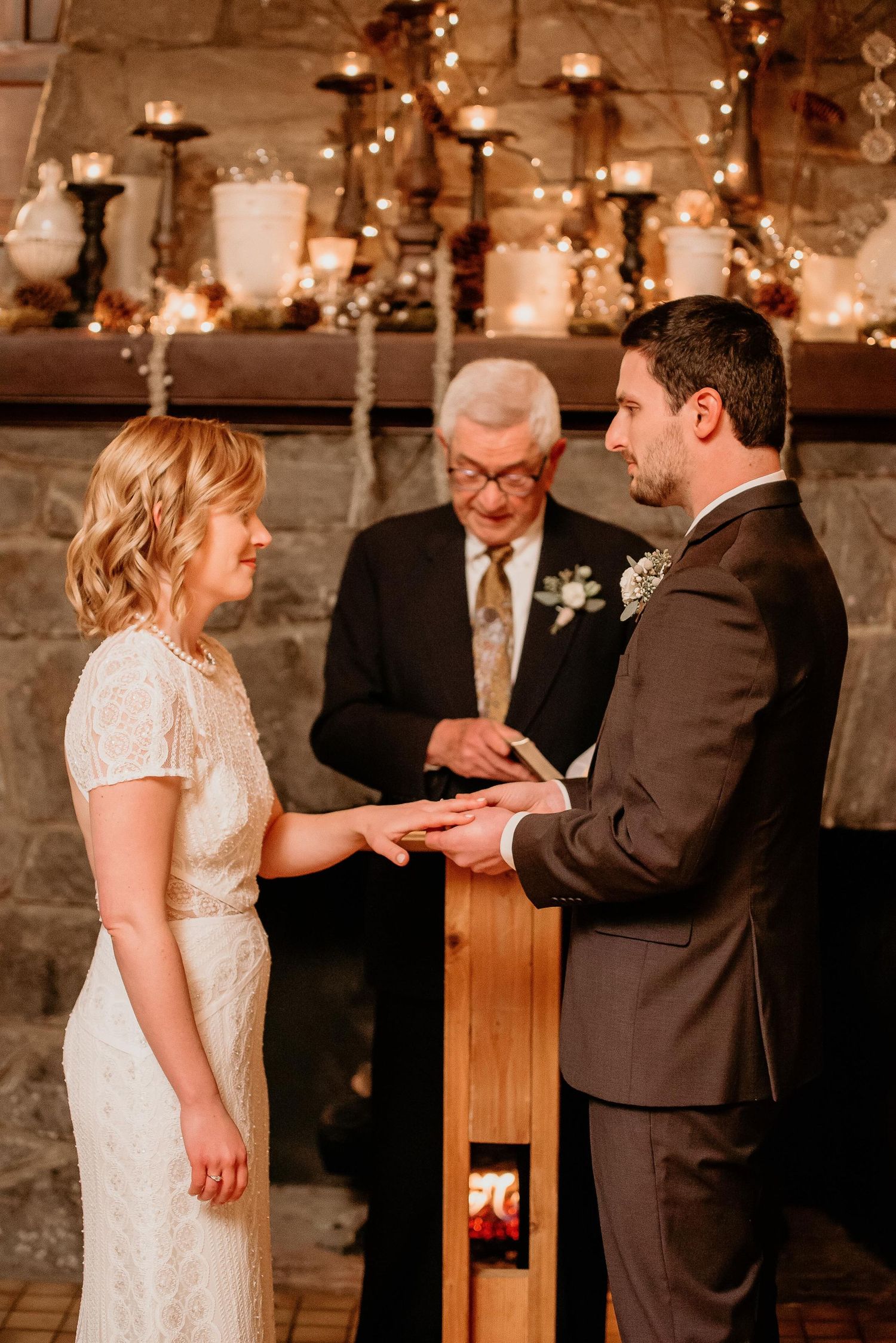 Bride and groom exchange rings at romantic indoor wedding ceremony with candlelit stone fireplace backdrop.