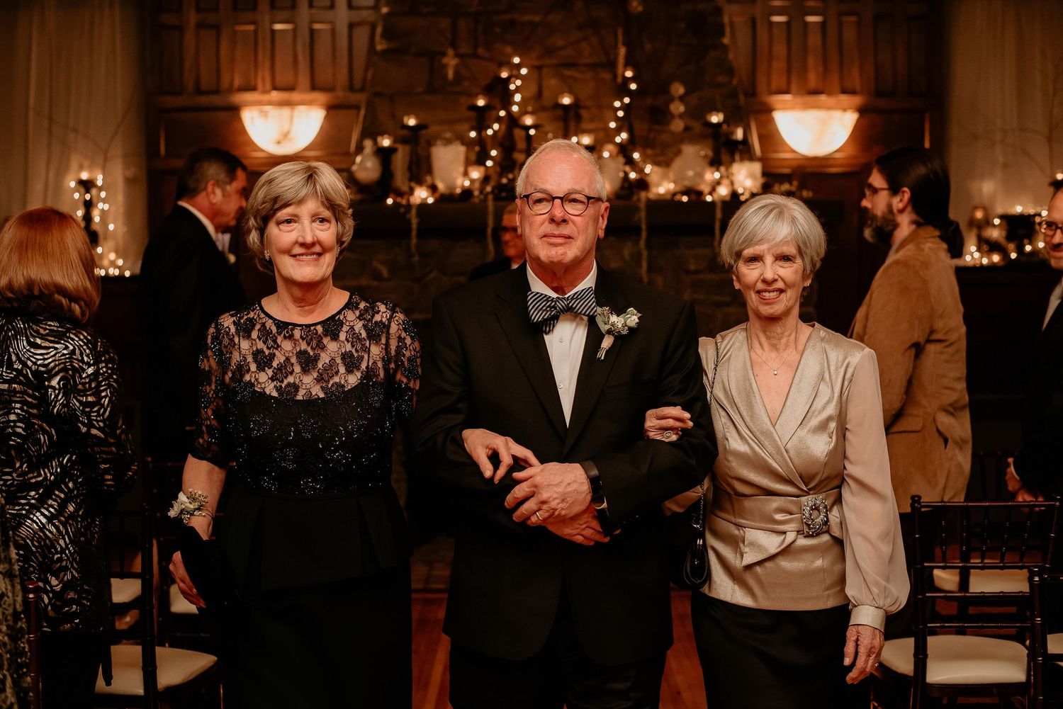 Elegant trio posing at formal wedding reception with fireplace and candlelit decor in background.