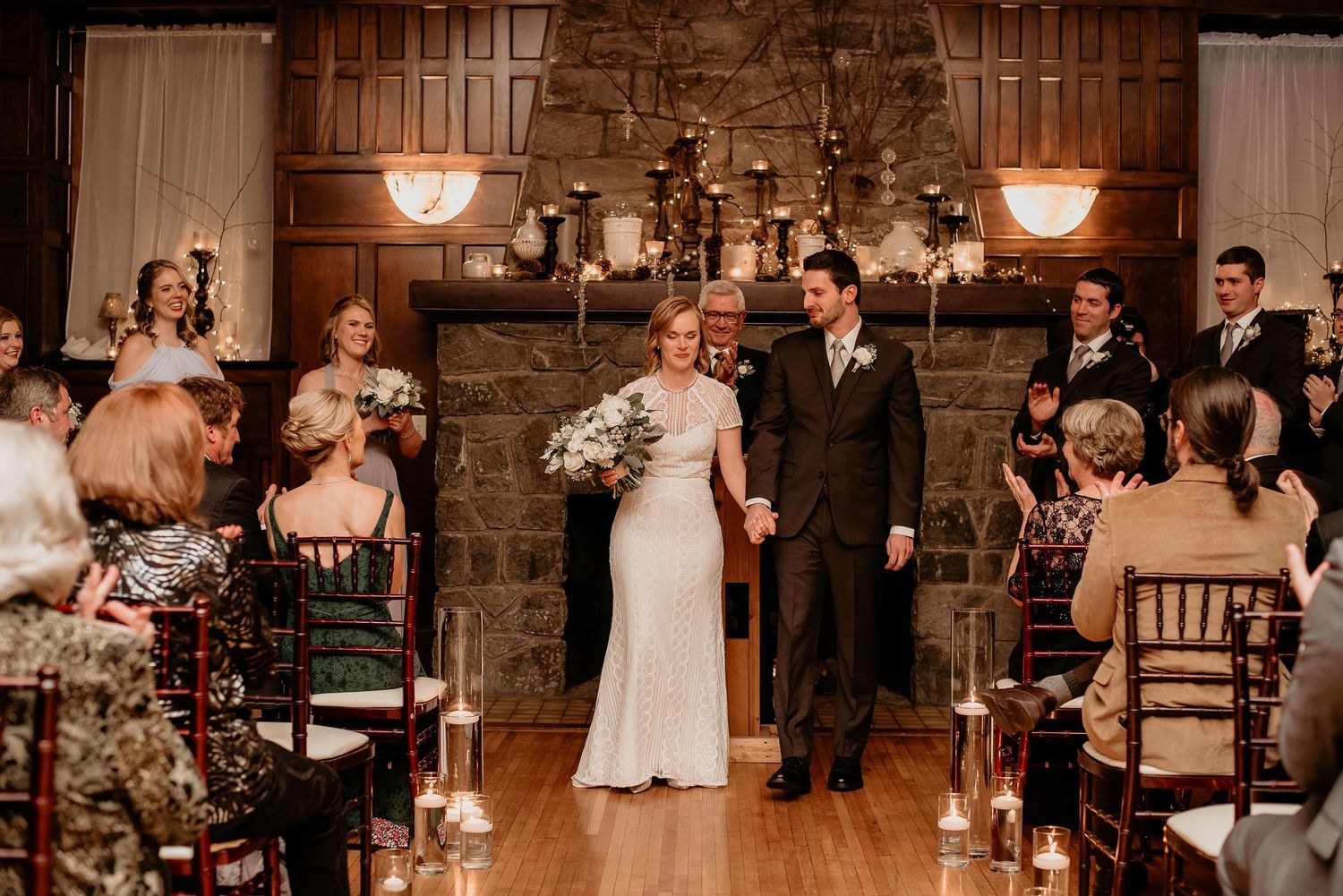 Bride and groom walk down the aisle at an elegant indoor wedding ceremony with candlelit chairs and stone fireplace.