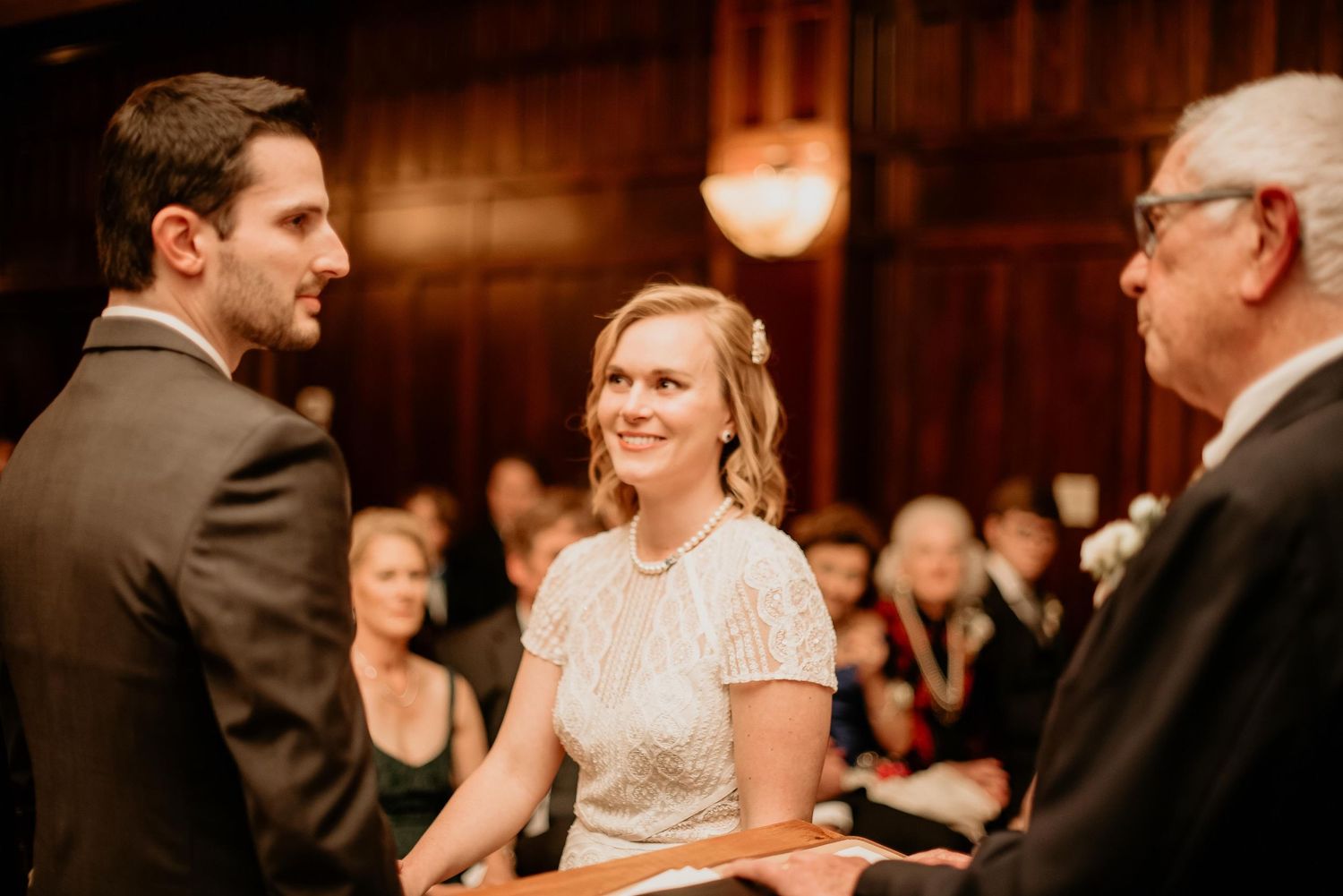 Bride and groom exchange vows at elegant indoor wedding ceremony with guests seated in wooden pews behind them.