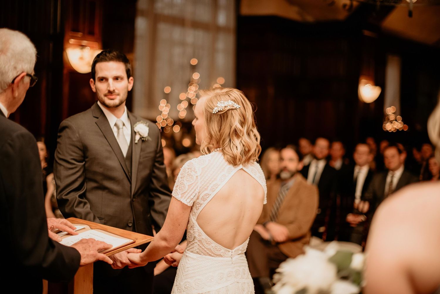 Groom in gray suit watches bride during wedding ceremony in warmly lit venue with open-back lace dress.