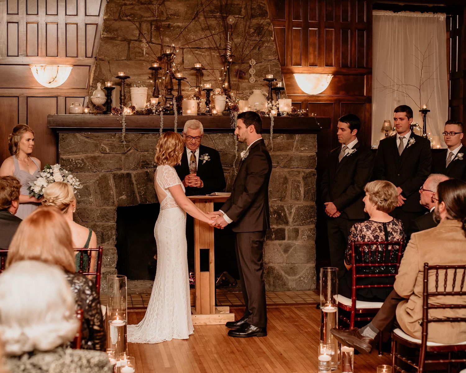 Couple exchanging vows at indoor wedding ceremony by stone fireplace with officiant and wedding party.