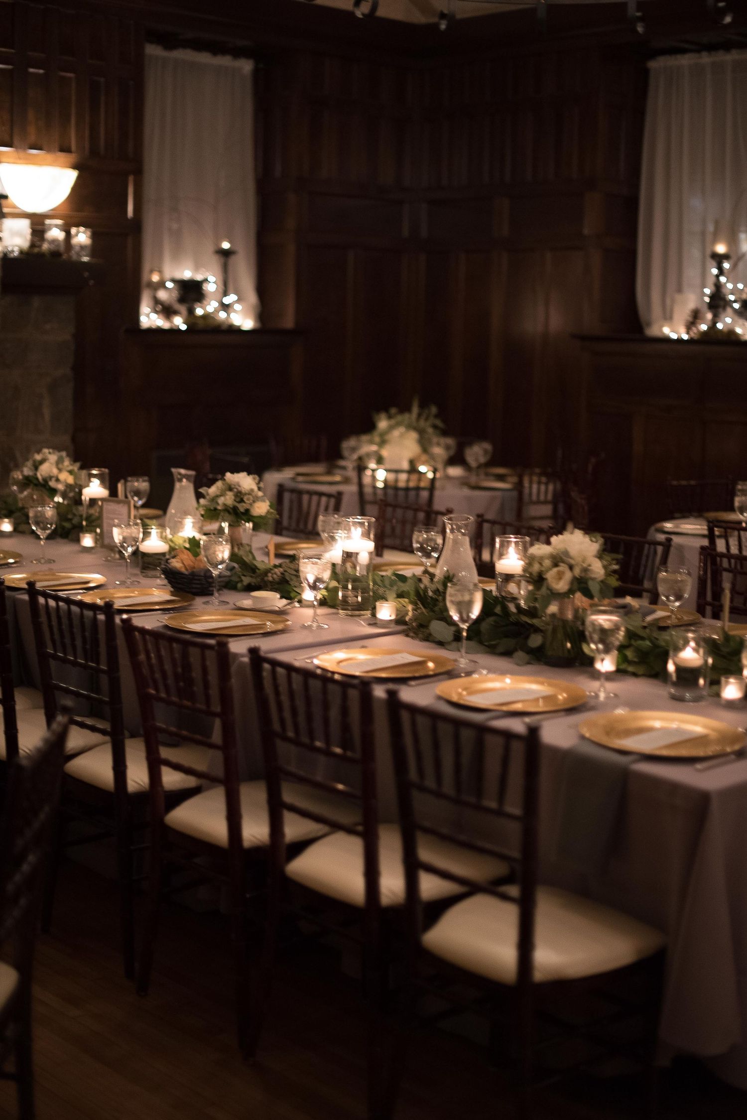 Elegant candlelit wedding reception with long tables, greenery garlands, gold chargers, and chiavari chairs in a dark wood venue.