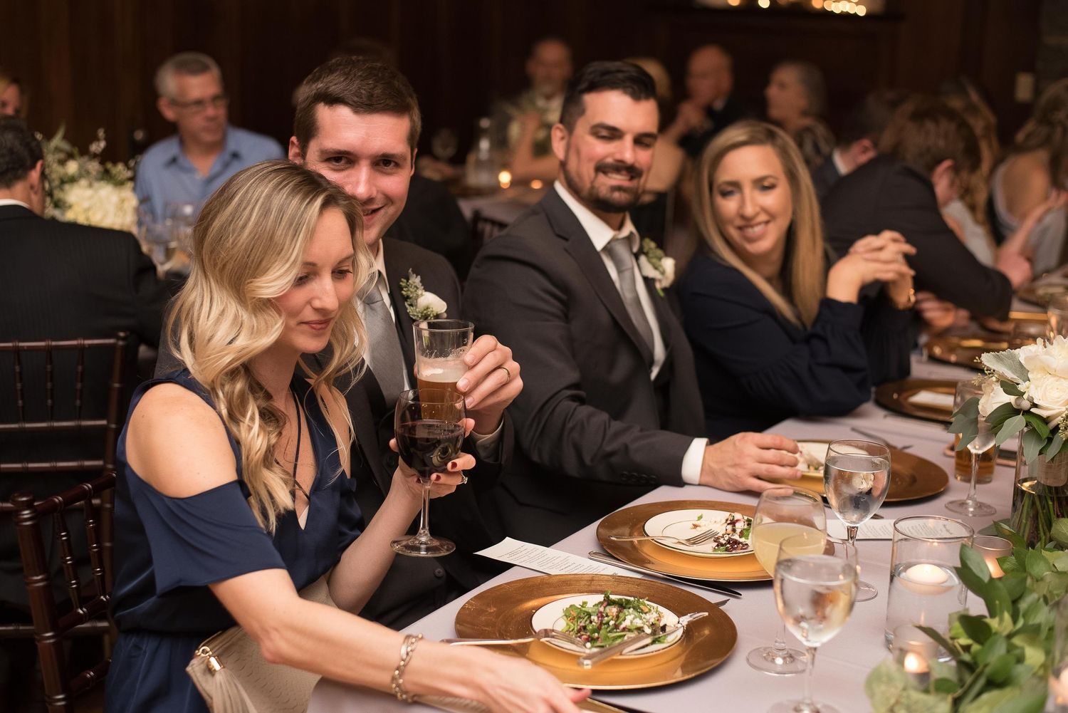 Guests smiling and raising glasses at an elegant wedding reception dinner table with floral centerpieces.