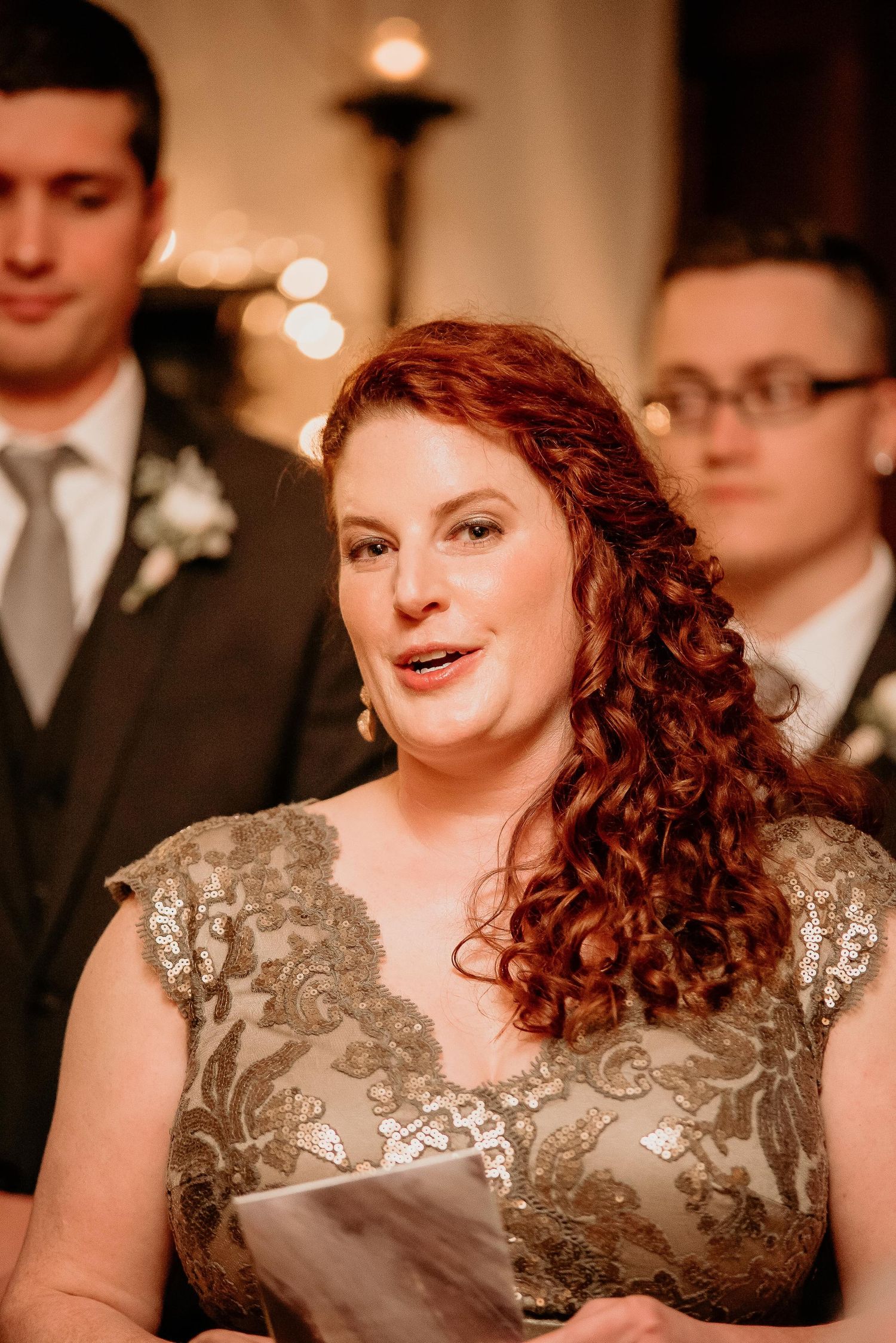 Red-haired woman in gold lace dress speaking at a wedding ceremony, holding a program.