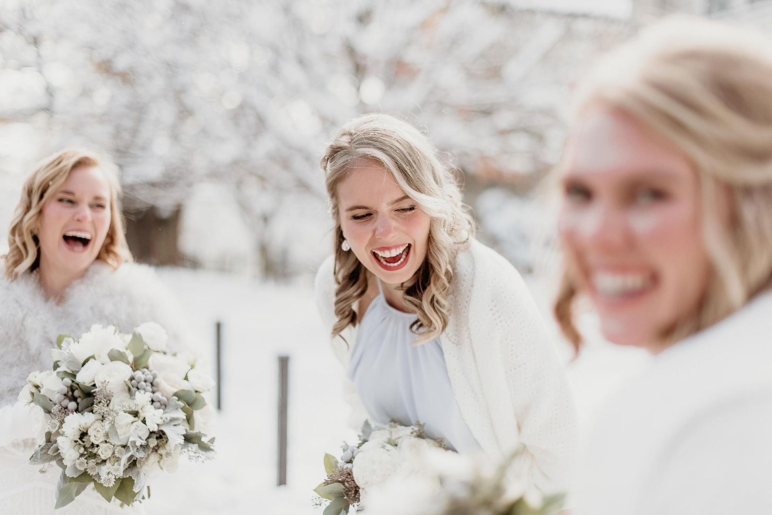 Three bridesmaids in white winter wedding attire laughing joyfully while holding white floral bouquets outdoors in snow.