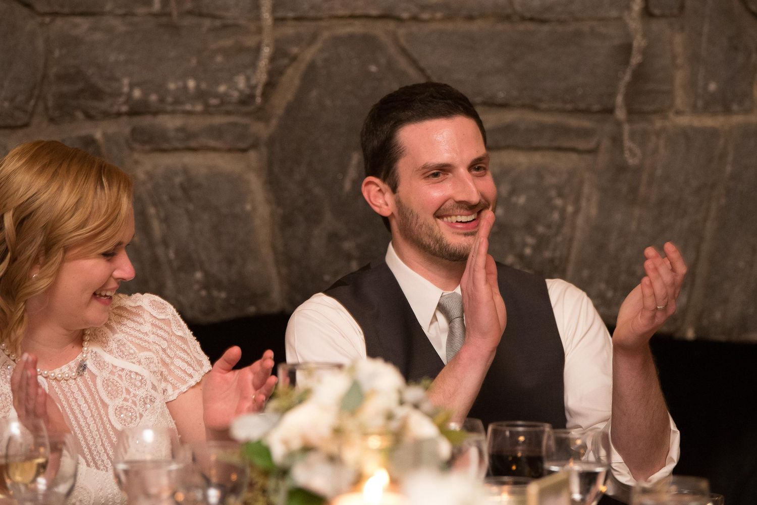 Man and woman clapping and smiling at a formal dinner event, seated at a decorated table with stone wall backdrop.