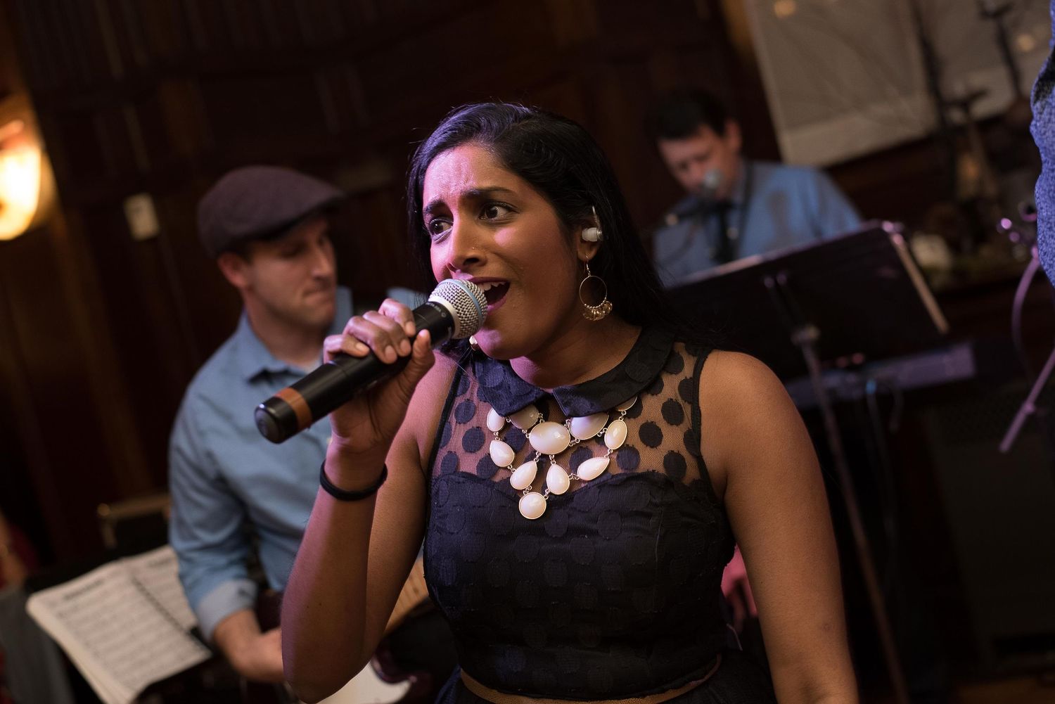 Woman singer performing live on stage with microphone, wearing black dress with gold necklace, band members visible behind her.