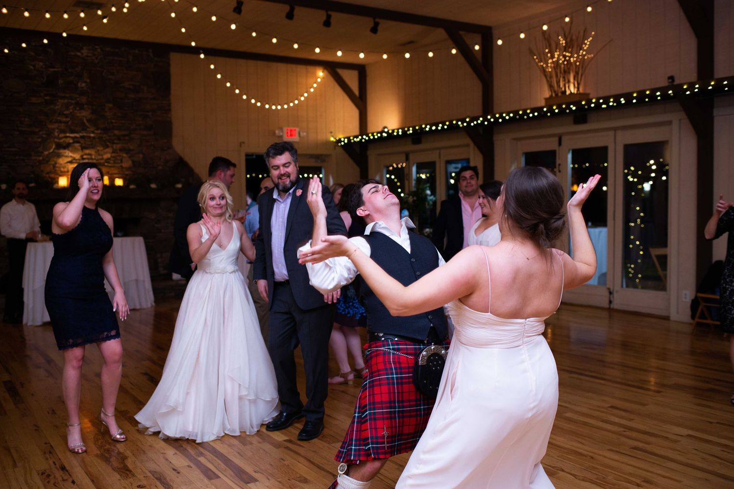 Wedding guests dancing joyfully in a rustic venue with string lights, man in red tartan kilt twirling bride on dance floor.