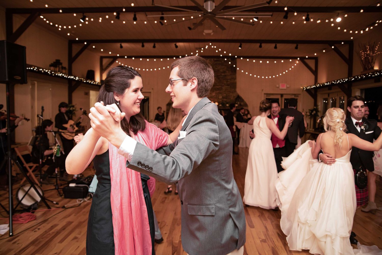 Couples dancing at a wedding reception in a warmly lit barn venue with string lights and a live band.