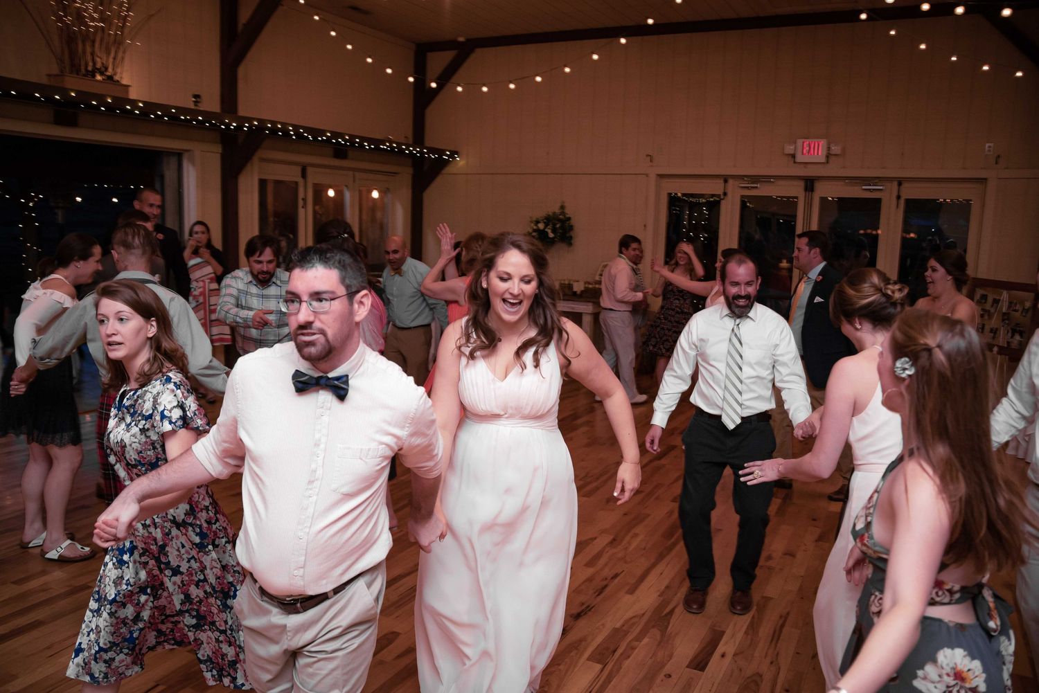 Bride and groom dance with guests at a lively wedding reception in a warmly lit rustic barn venue.