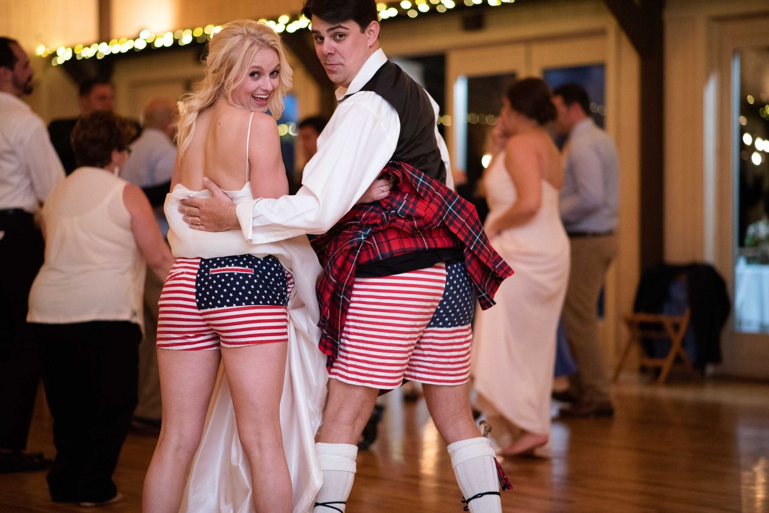Couple dancing at wedding reception wearing matching American flag shorts and Scottish kilt attire on dance floor.