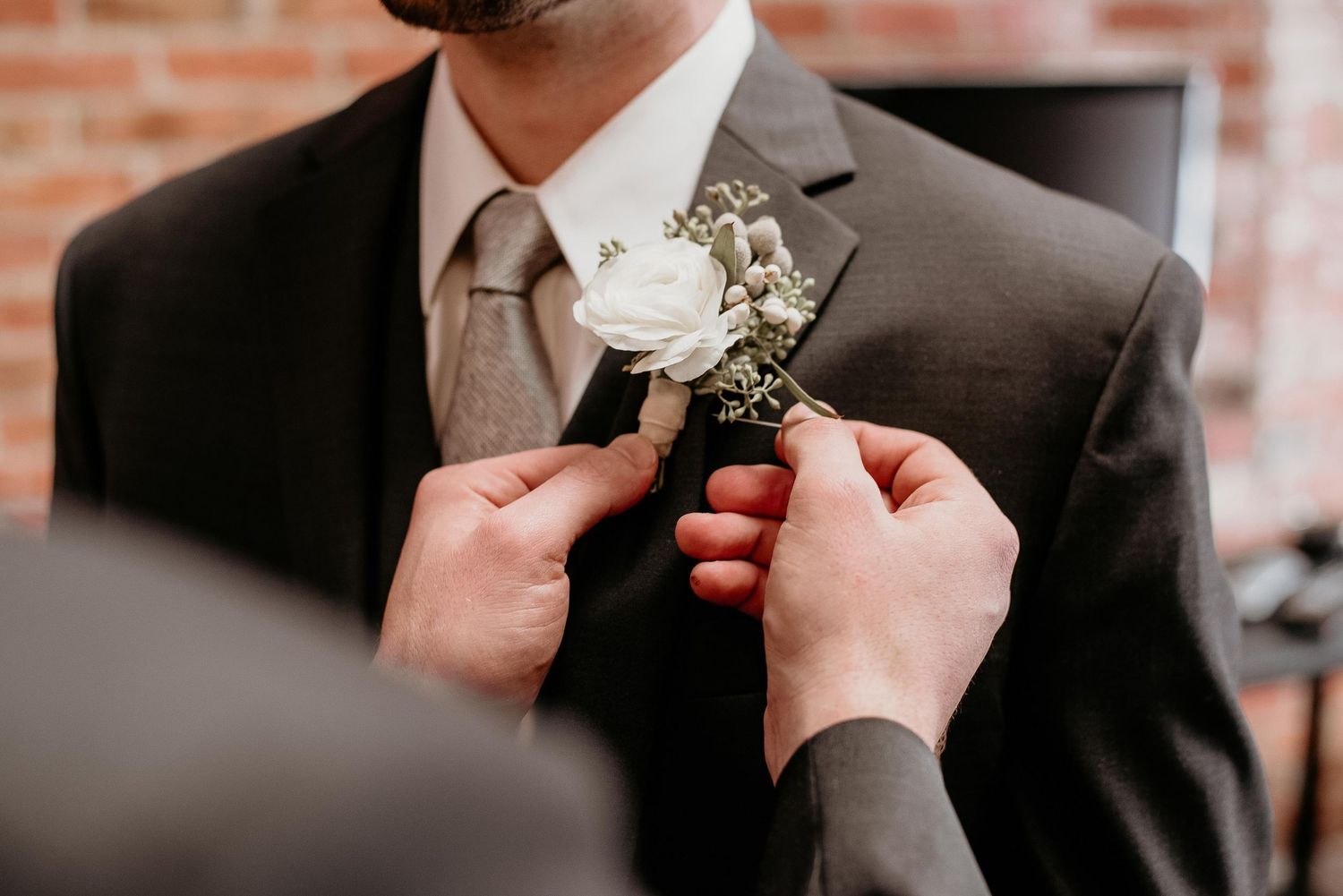 Hands pinning a white rose boutonniere on a groom's dark suit lapel at a wedding.