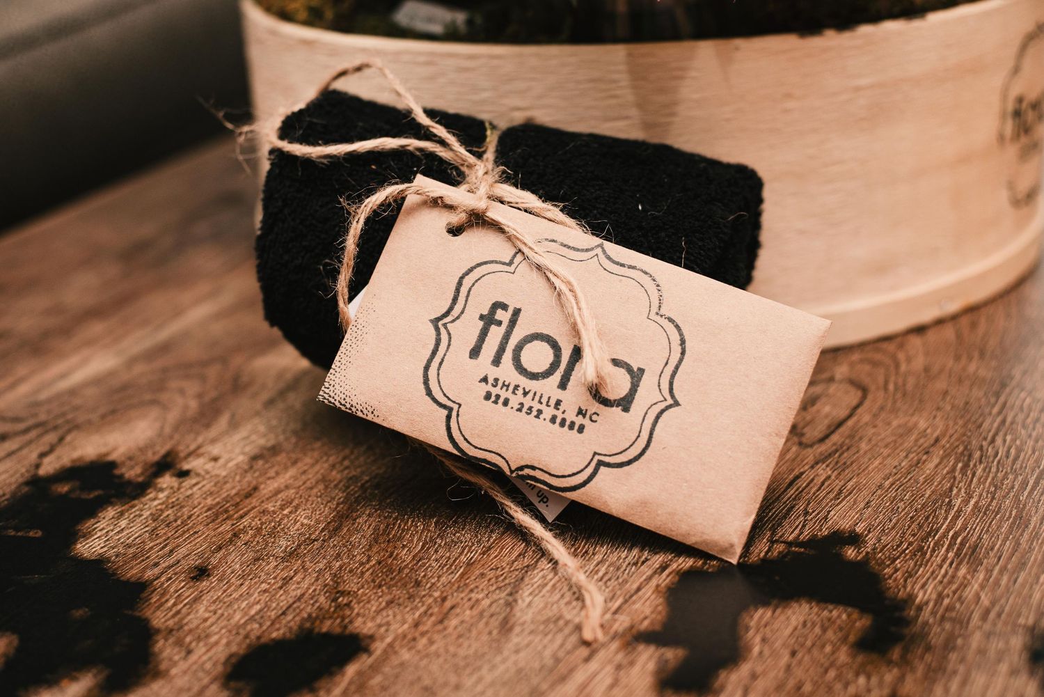 Flora branded gift tag tied with twine on a folded black cloth item displayed on a rustic wooden surface.