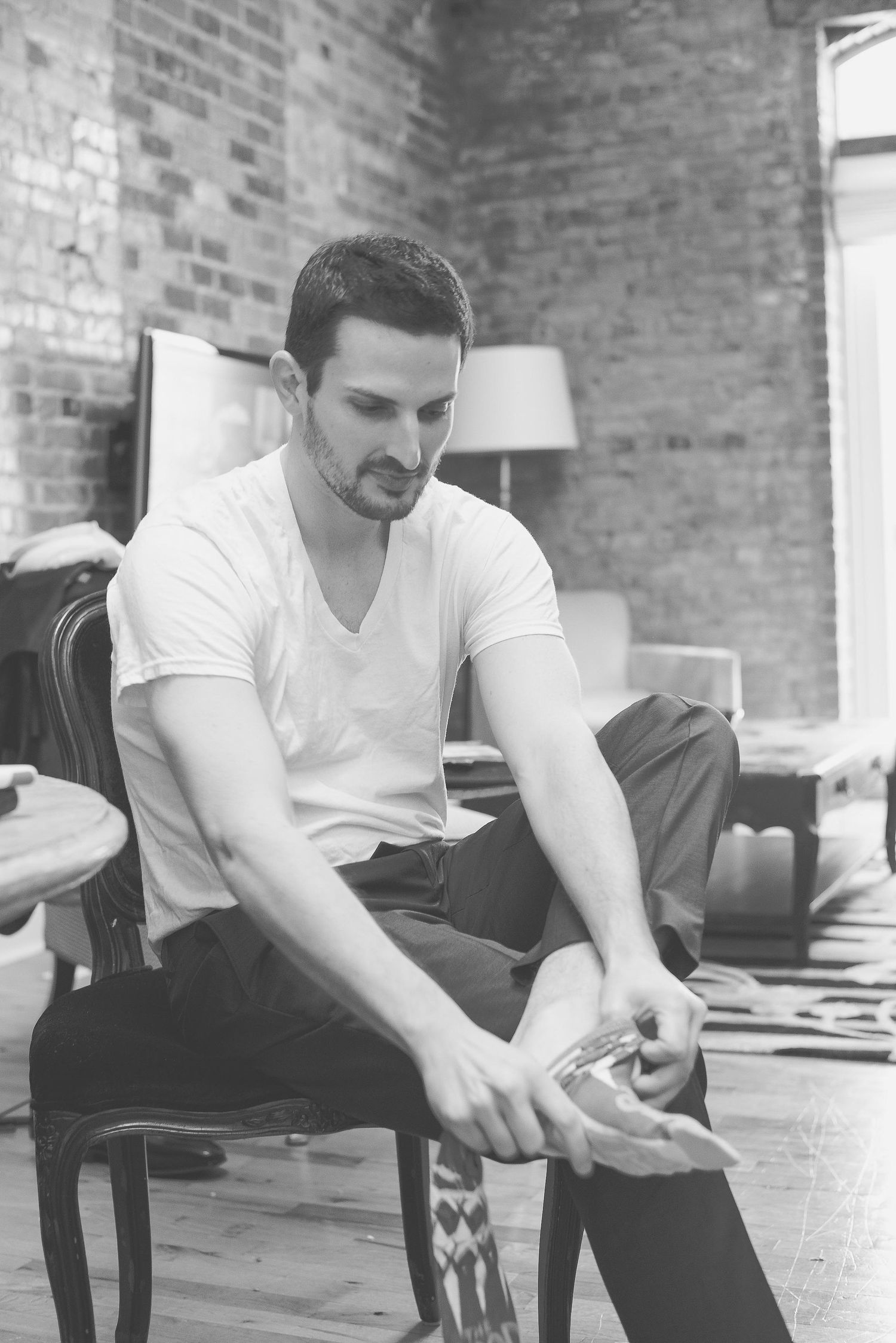 Black and white photo of a young man in white t-shirt sitting on chair, looking down at his hands indoors.