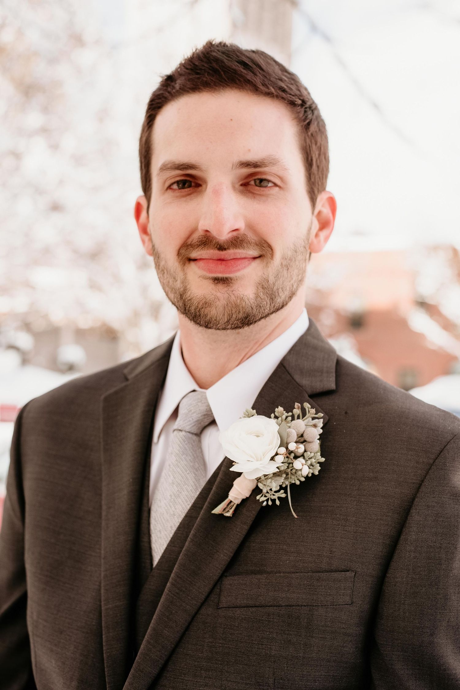Smiling groom in gray suit with white rose boutonniere poses for wedding portrait outdoors.