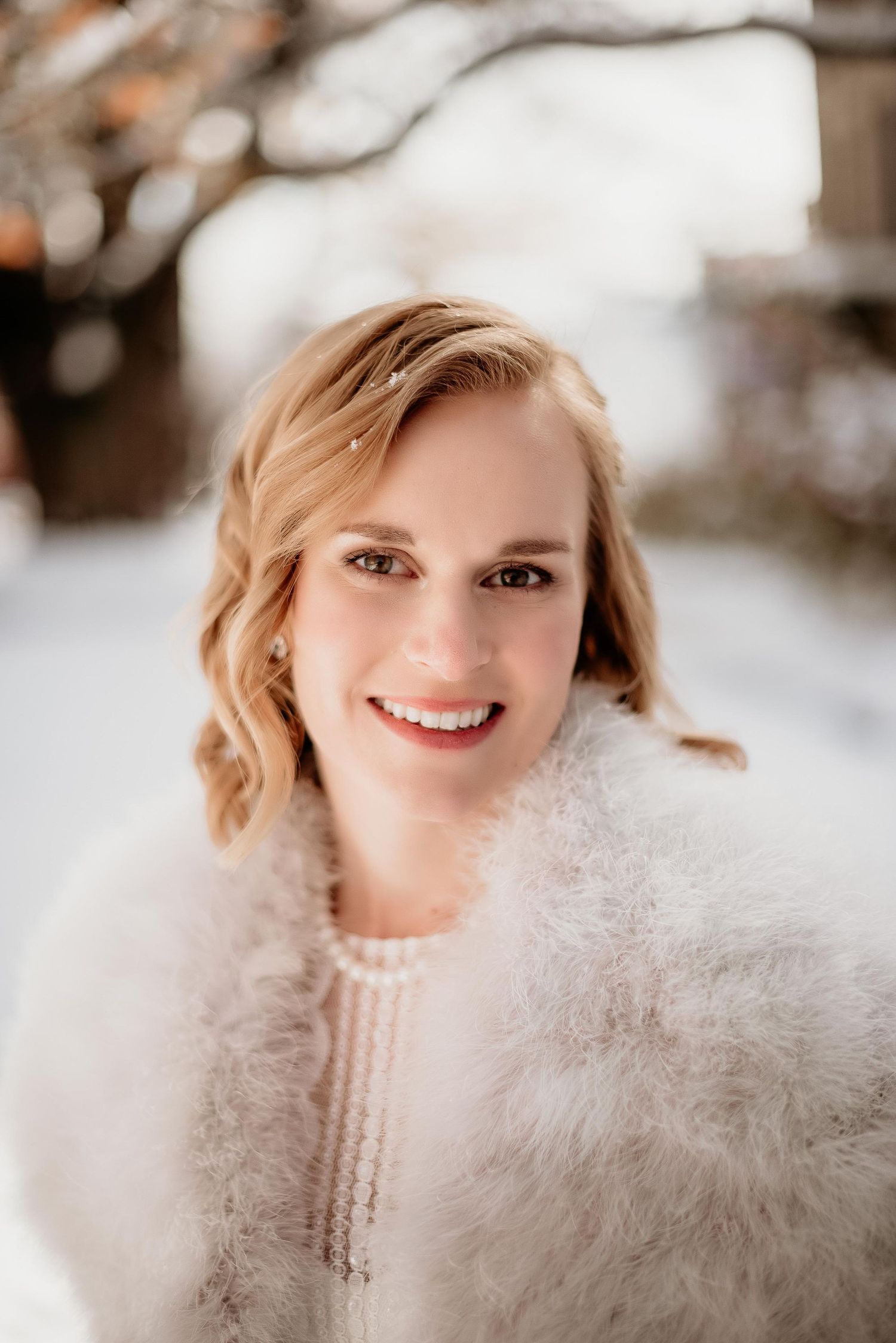 Smiling blonde woman in white fur wrap and pearl necklace posing outdoors in a snowy winter setting.