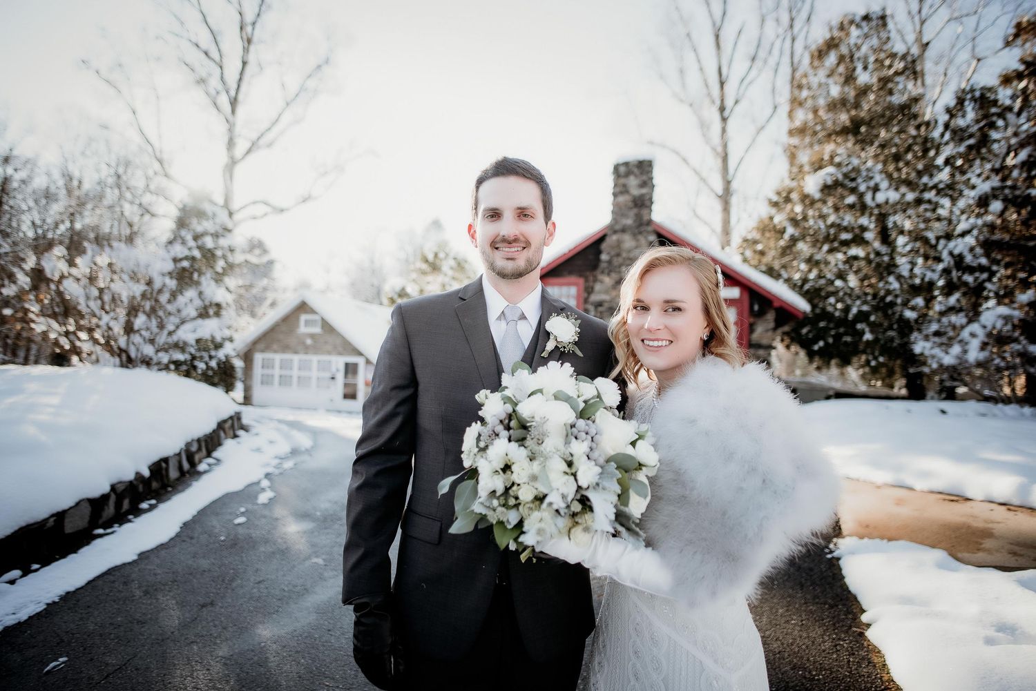 Bride and groom smiling together in winter snow, bride holding white floral bouquet with fur wrap, red cabin background.