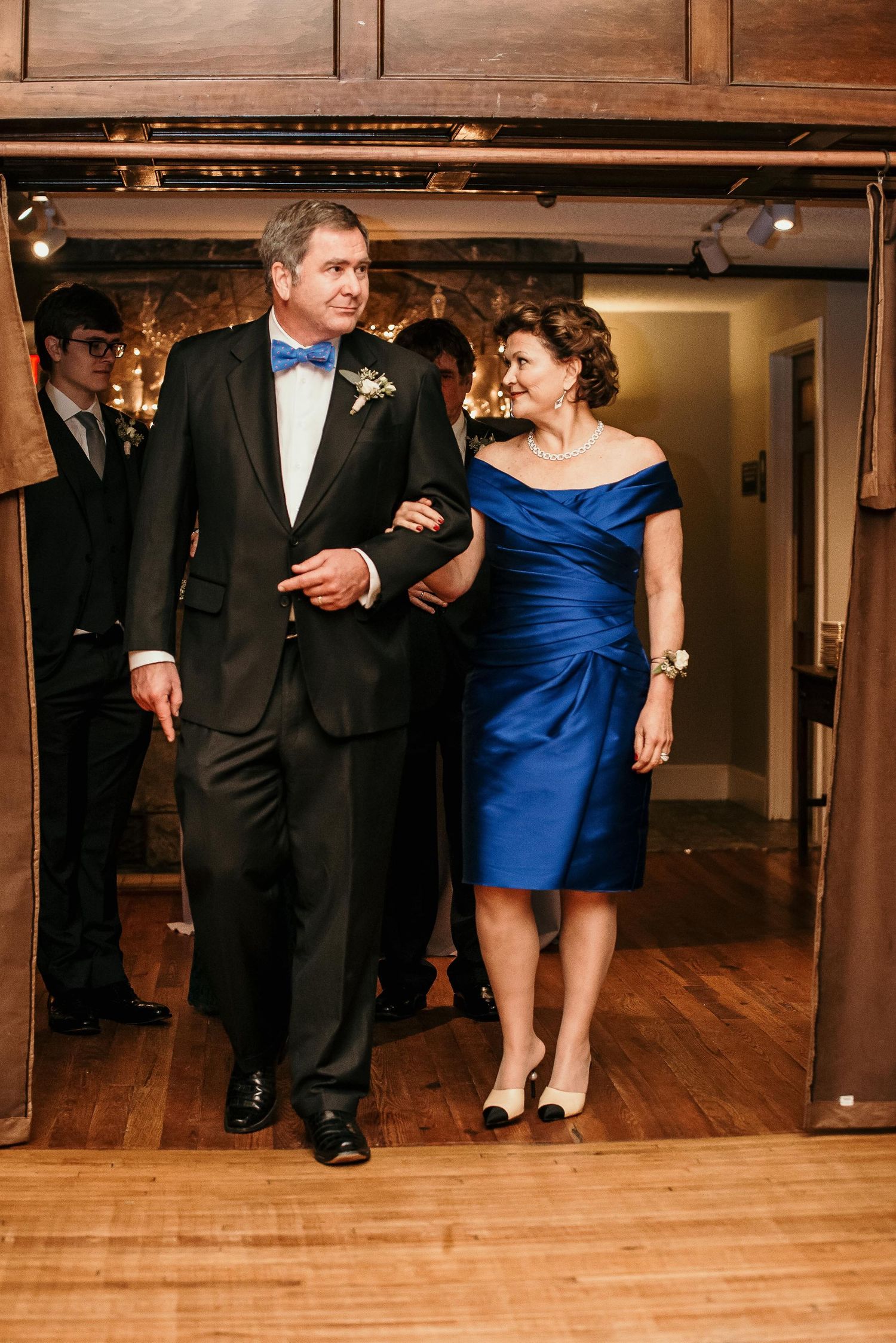 Groom in black tuxedo with blue bow tie escorts woman in royal blue off-shoulder dress at wedding reception.