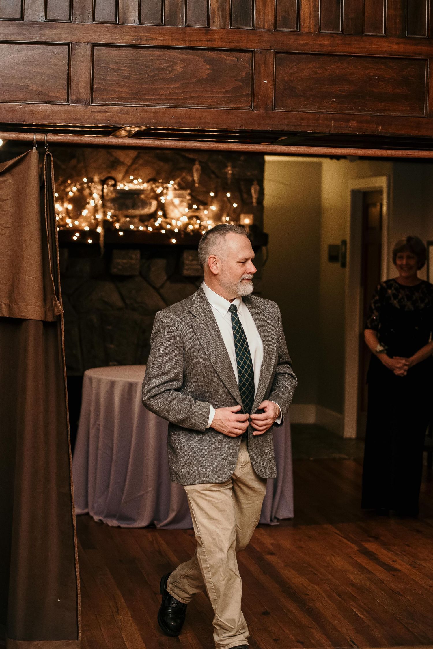 A man in a gray blazer and striped tie walks through a warmly lit rustic venue with string lights and wooden decor.