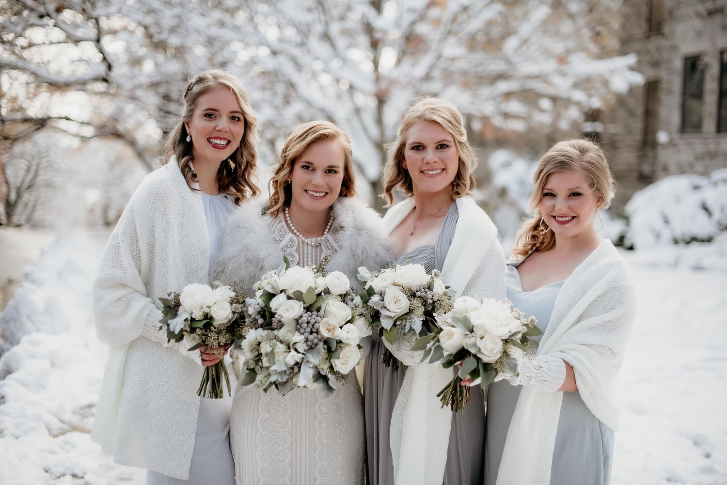 Four bridesmaids in gray dresses and white wraps holding white floral bouquets posing outdoors in a snowy winter wedding.