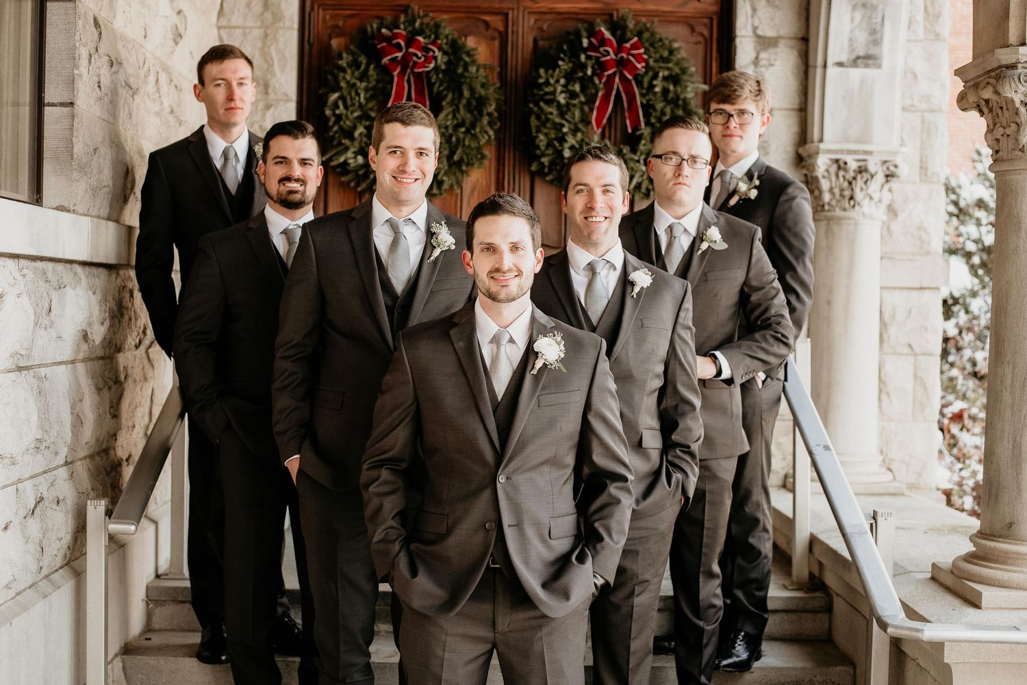 Groom and groomsmen in matching brown suits pose on stone steps with holiday wreaths at a winter wedding.