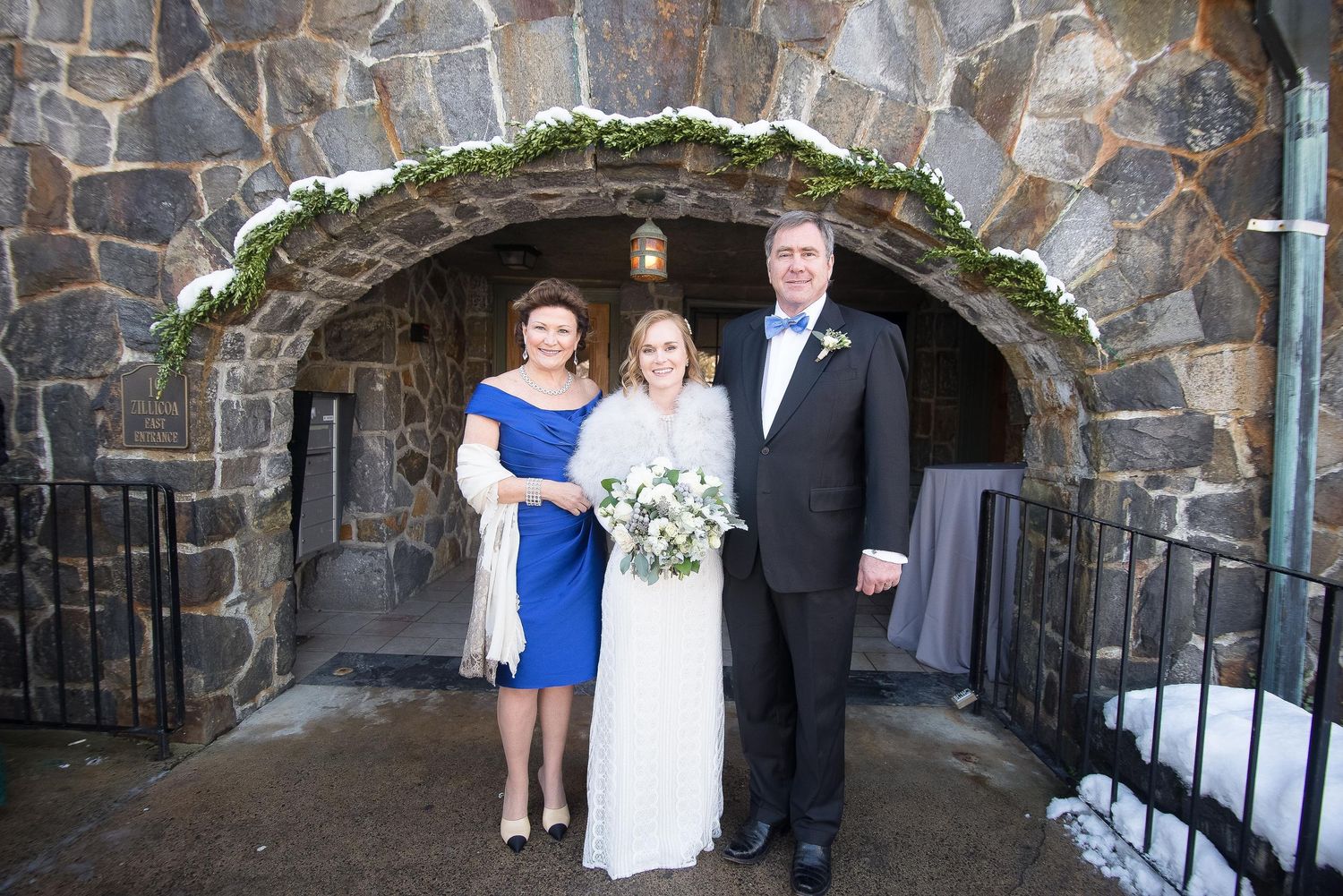 Bride in white gown poses with parents in front of stone arch entrance at winter wedding venue.