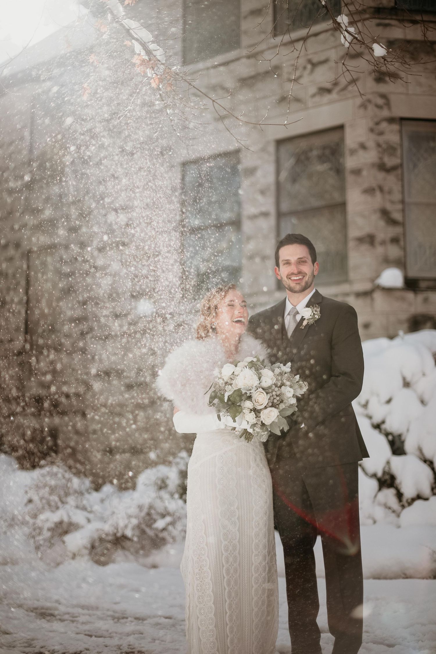 Bride and groom laughing in snowfall outside stone building, winter wedding portrait with white bouquet.