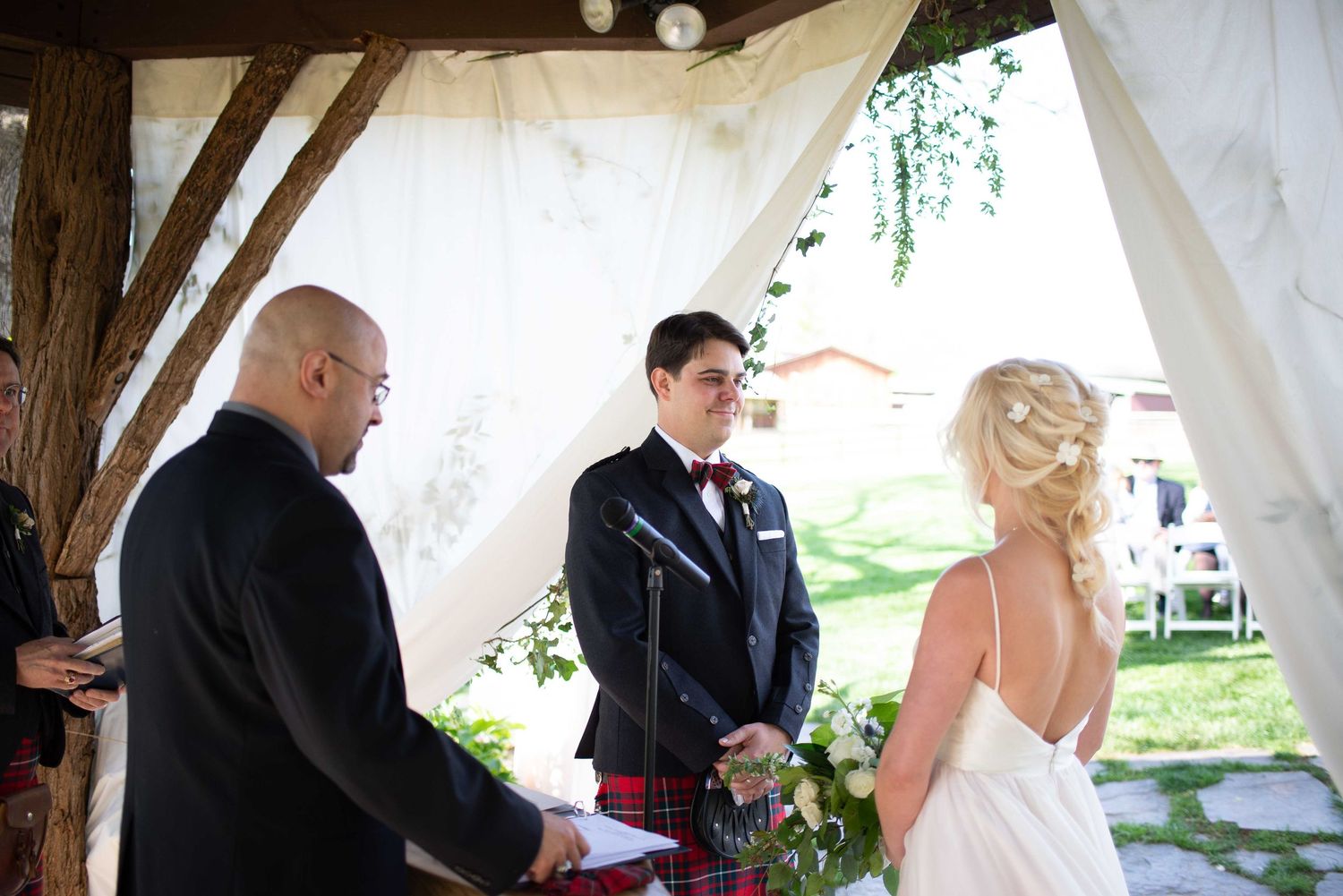Groom in kilt and bow tie stands with bride in white dress during outdoor wedding ceremony under draped canopy.