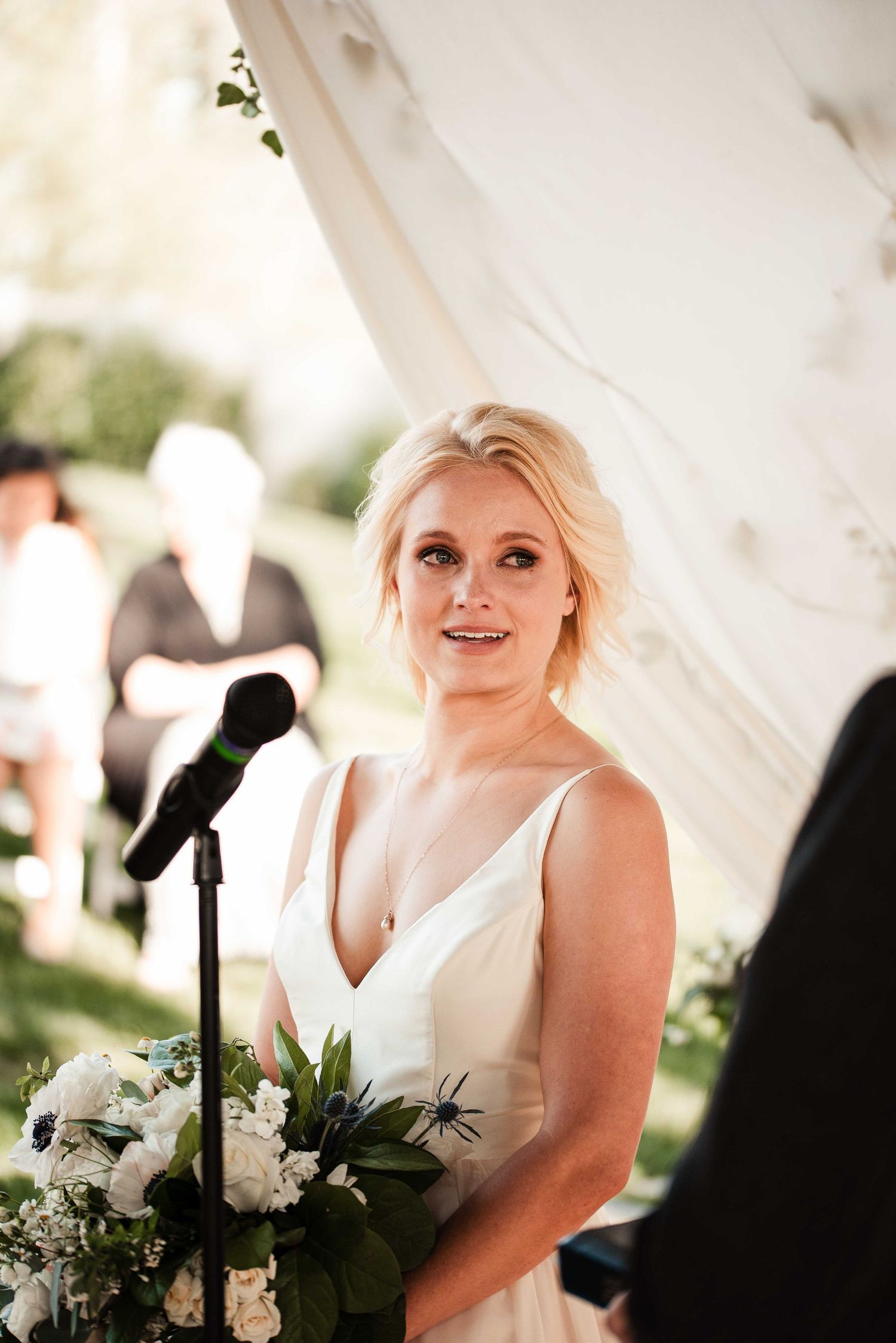 Blonde bride in white V-neck gown holds floral bouquet at outdoor wedding ceremony near microphone.