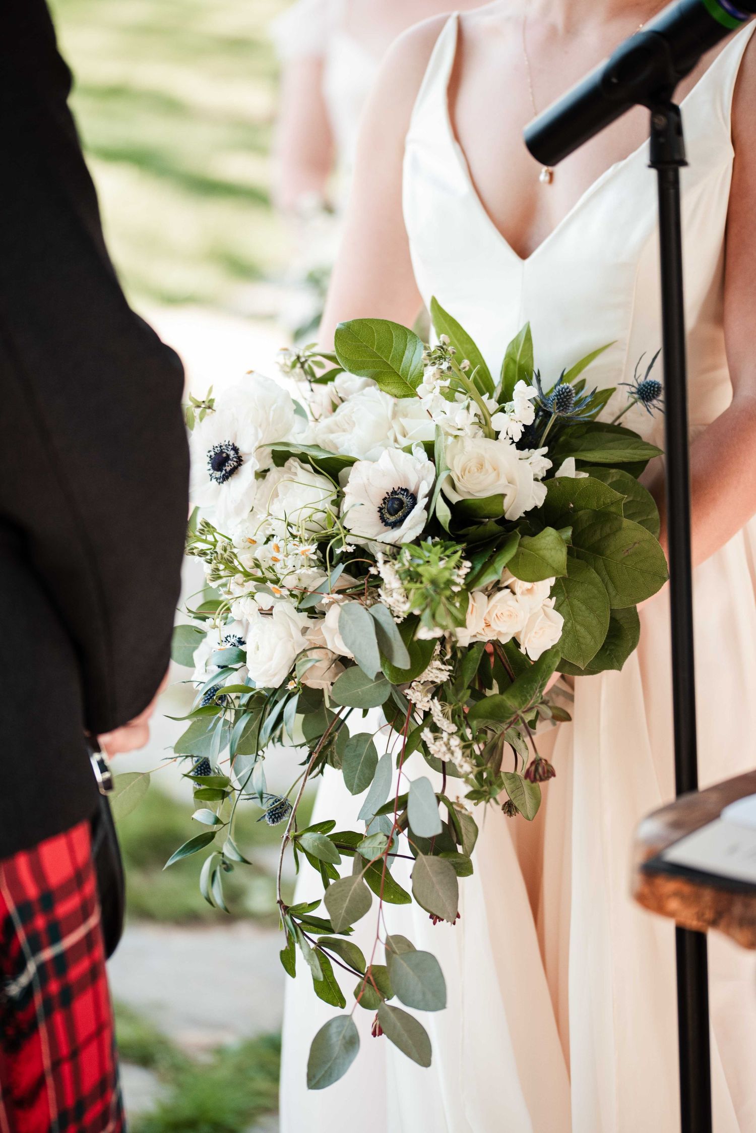 Bride holding lush cascading wedding bouquet with white anemones, roses, and greenery at outdoor ceremony.