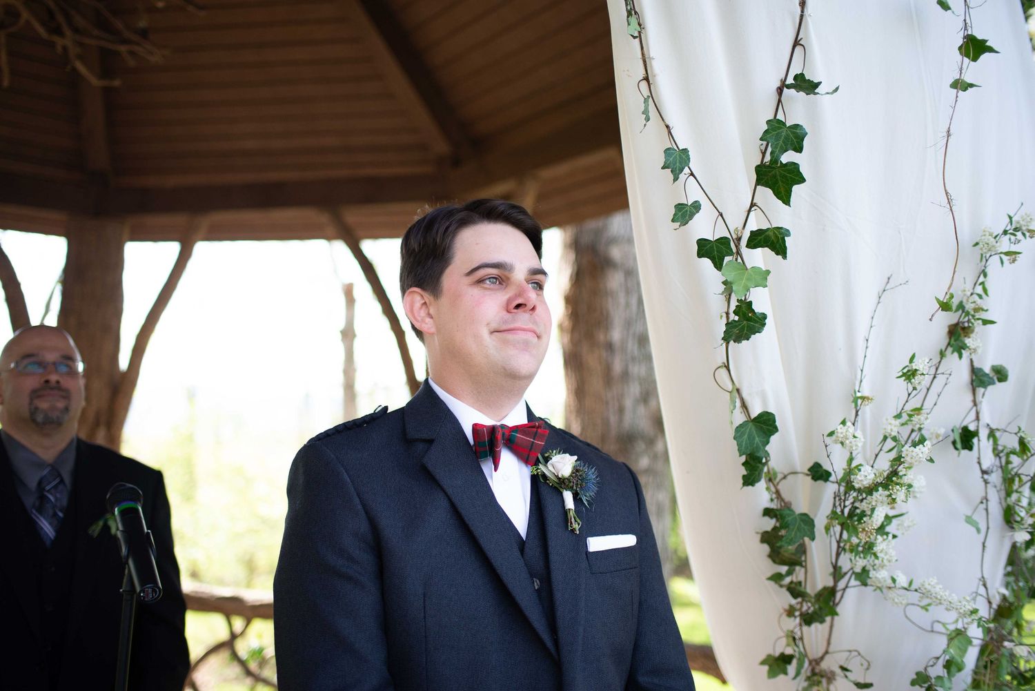 Groom in navy suit and burgundy bow tie smiling at altar during outdoor wedding ceremony.