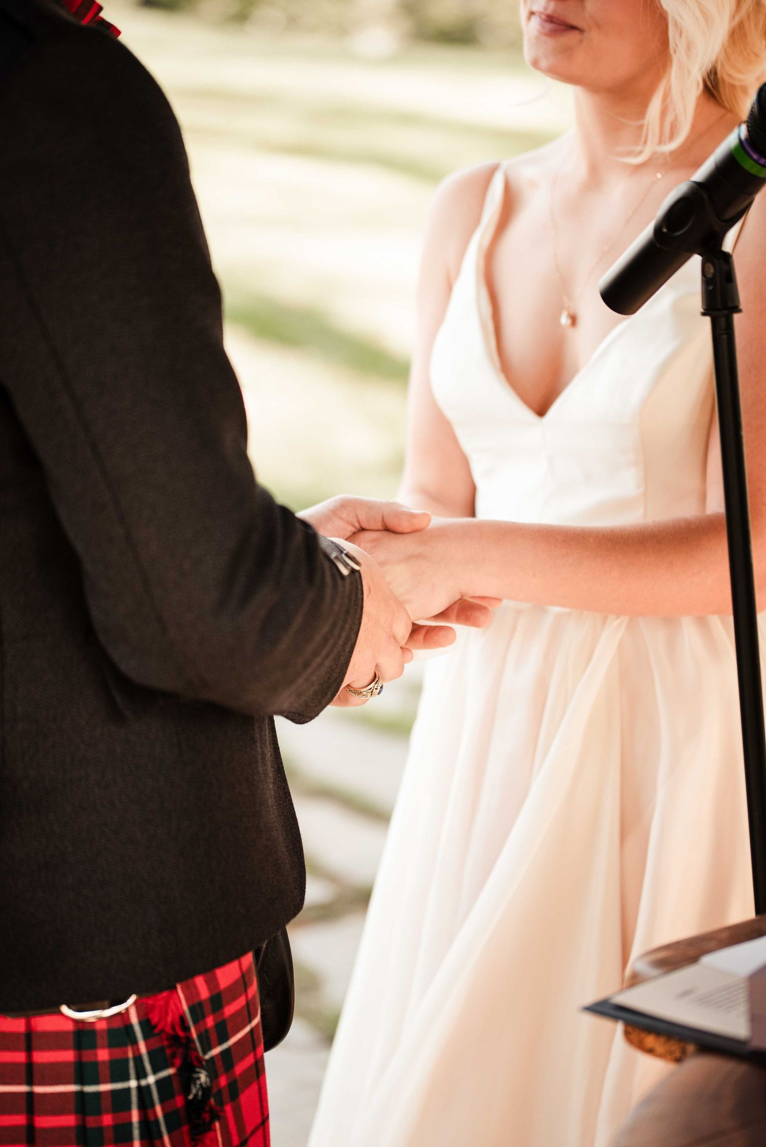 Bride and groom holding hands during wedding ceremony, groom wearing traditional red tartan kilt and black jacket.