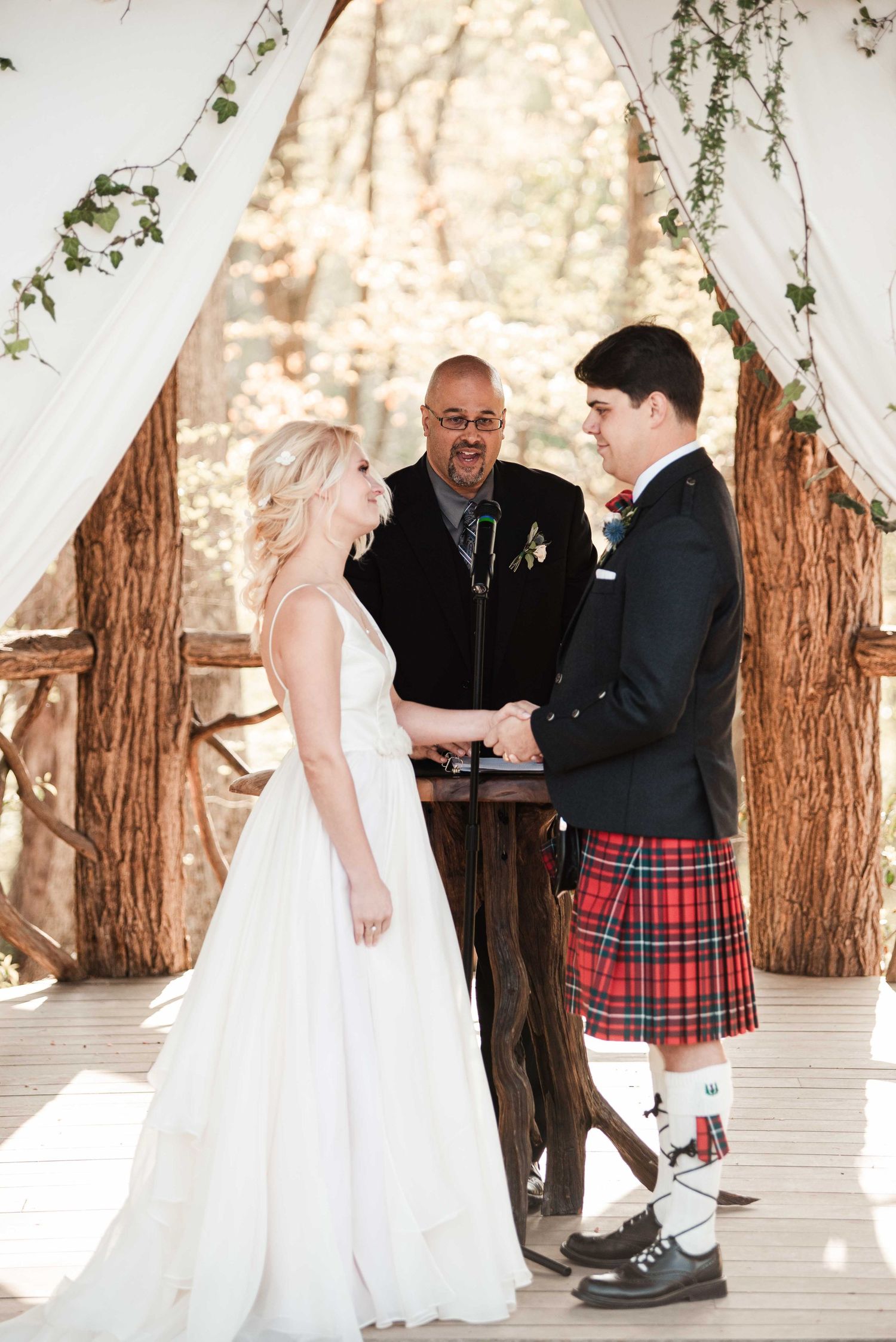 Bride and groom exchange vows outdoors, groom wearing red tartan kilt, officiant standing behind wooden podium.