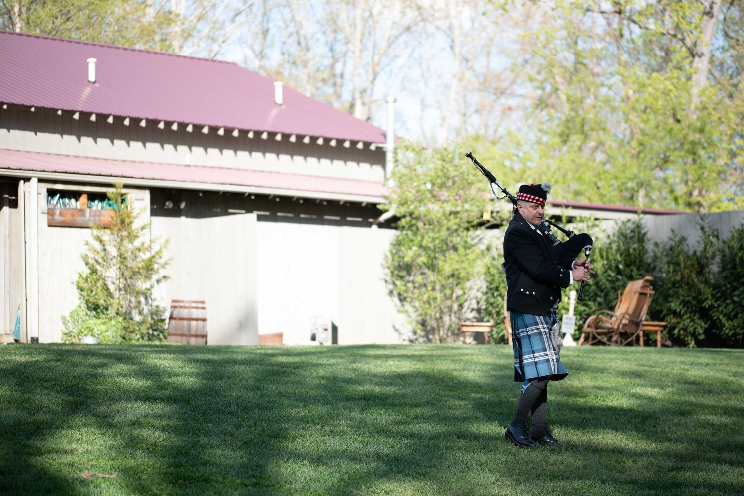 Bagpiper in traditional Scottish kilt performs outdoors on green lawn beside a building on a sunny day.
