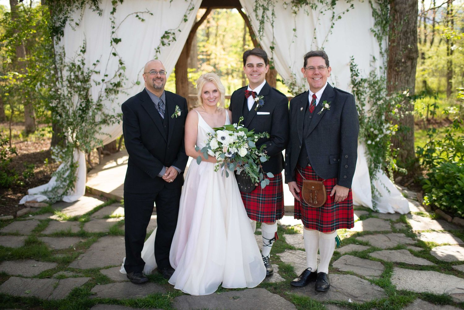 Wedding party poses outdoors at floral arch, bride in white gown with bouquet, groom in red tartan kilt, two men in suits.