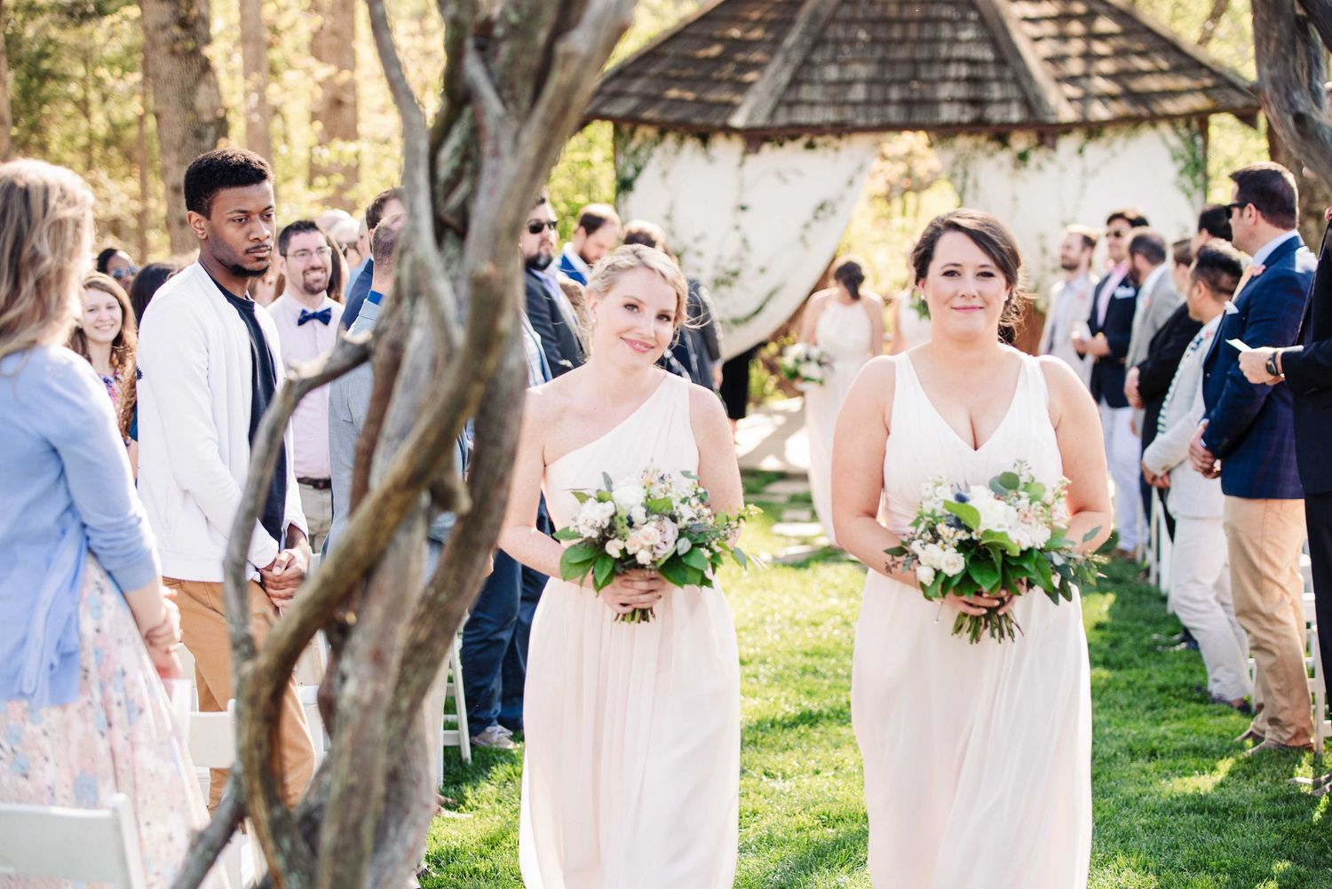 Two bridesmaids in blush gowns walk down outdoor wedding aisle holding white bouquets, guests seated on both sides.
