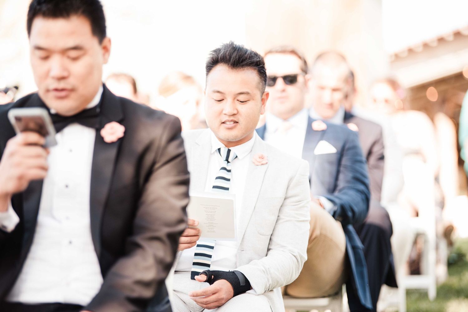 Men in formal suits at an outdoor wedding ceremony, one reading a program.