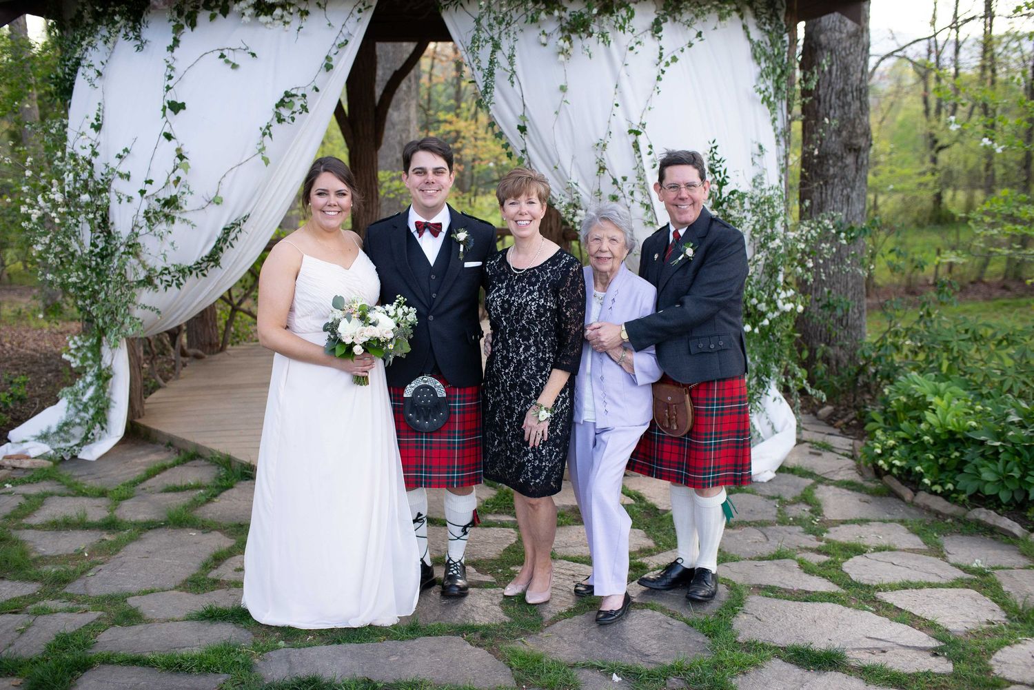 Wedding party in kilts and bridal gown pose under floral arch at outdoor ceremony.