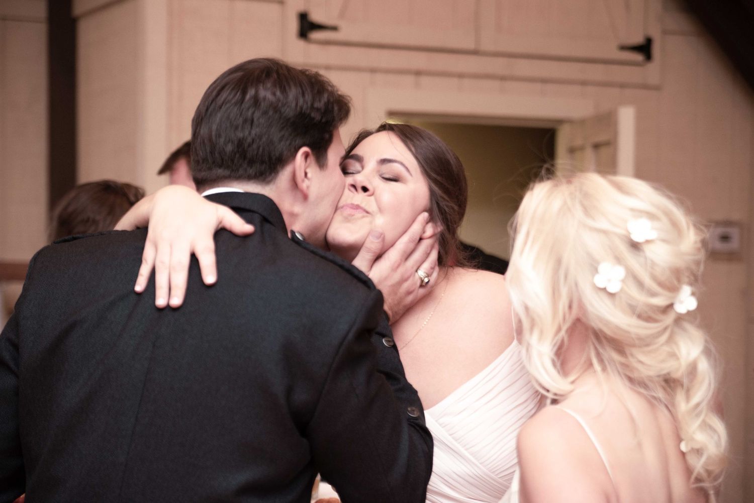 Groom kisses bride on cheek at wedding reception while blonde bridesmaid stands nearby.