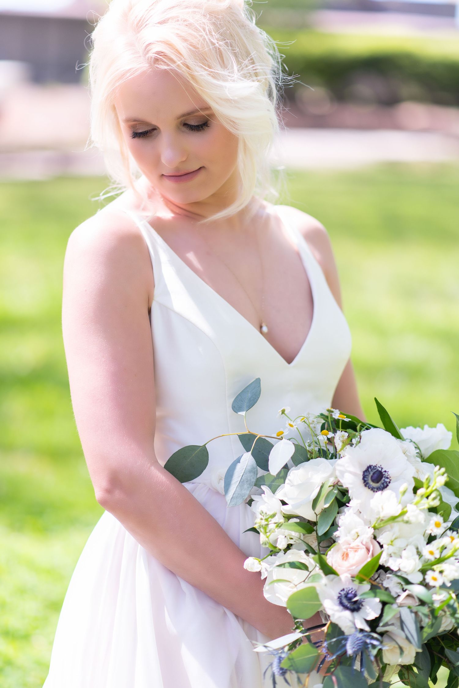 Elegant blonde bride in V-neck gown holding white anemone and eucalyptus bouquet.