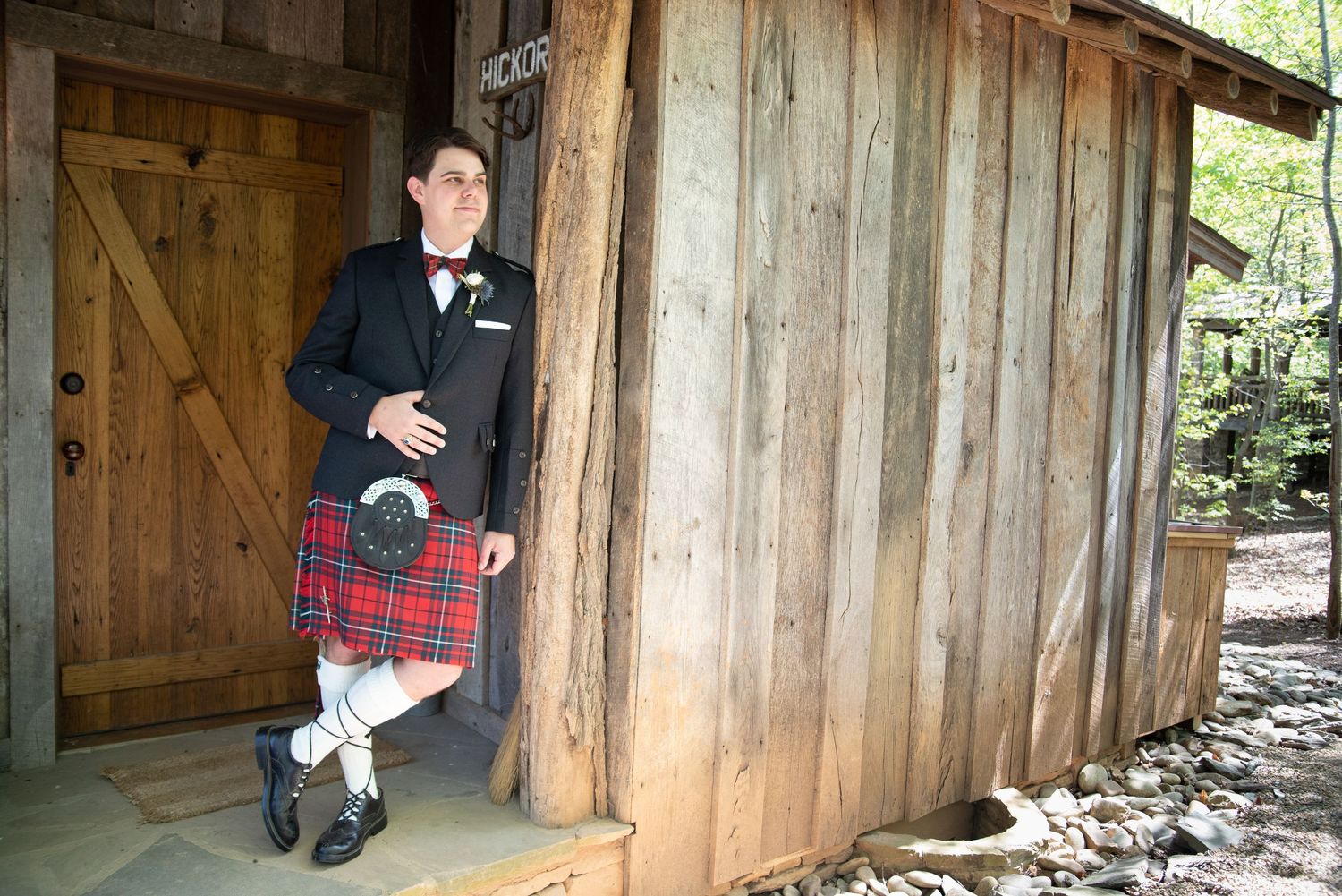 A groom wearing a traditional red tartan kilt and black jacket leaning against a rustic wooden barn.