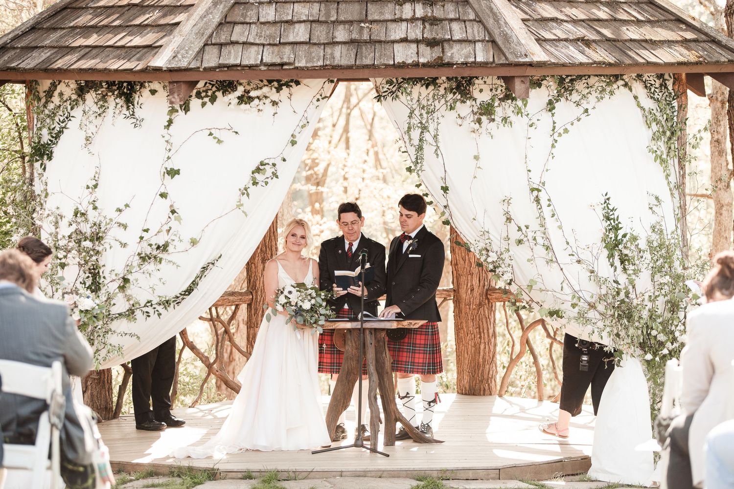 Bride and groom exchange vows under a rustic wooden gazebo adorned with white draping and greenery at an outdoor wedding.