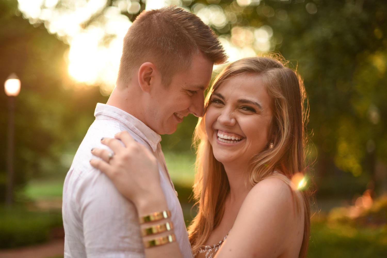 A smiling couple embraces outdoors at golden hour, with warm sunlight filtering through trees in a romantic park setting.