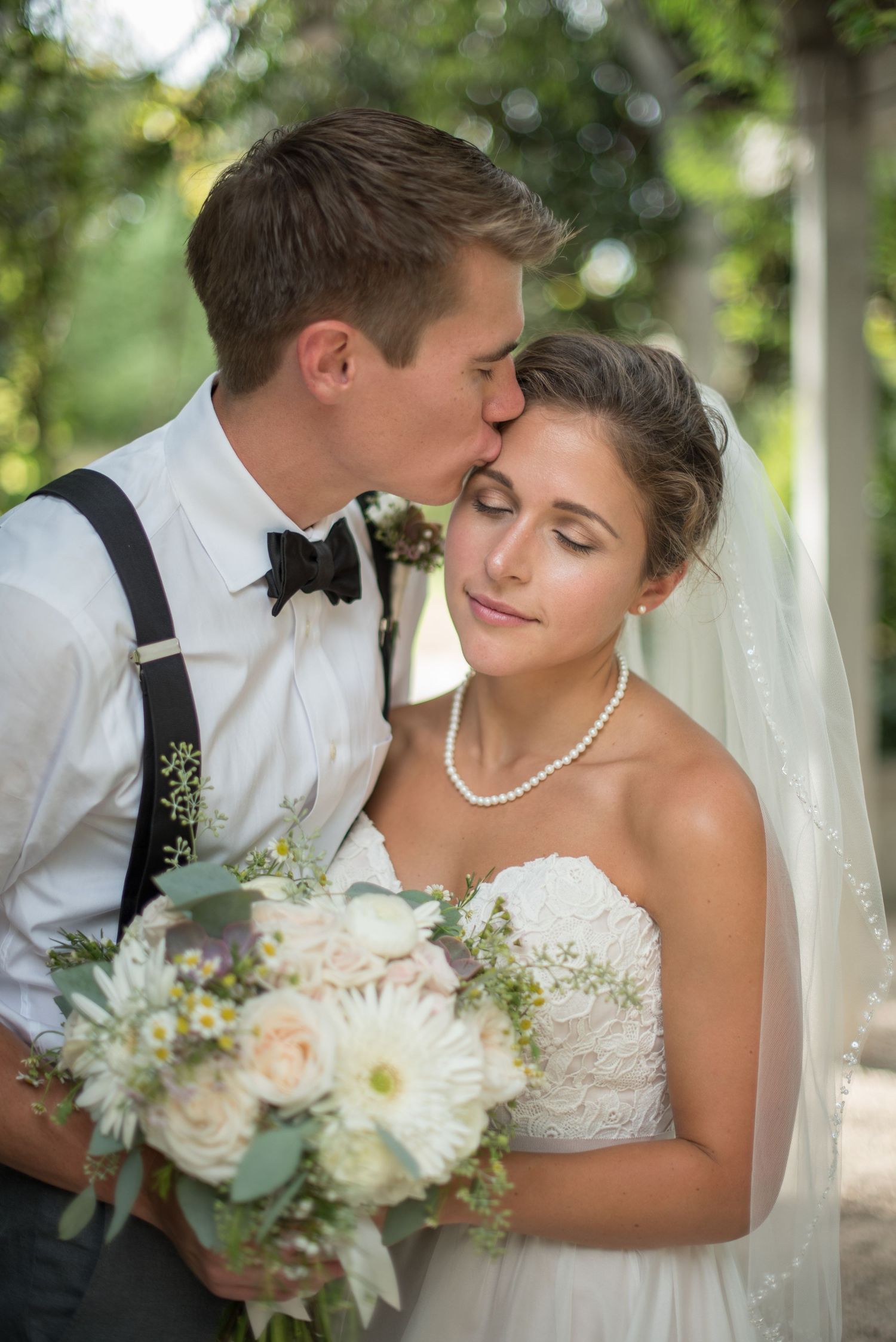 Groom kissing bride on forehead while she holds white floral bouquet outdoors.