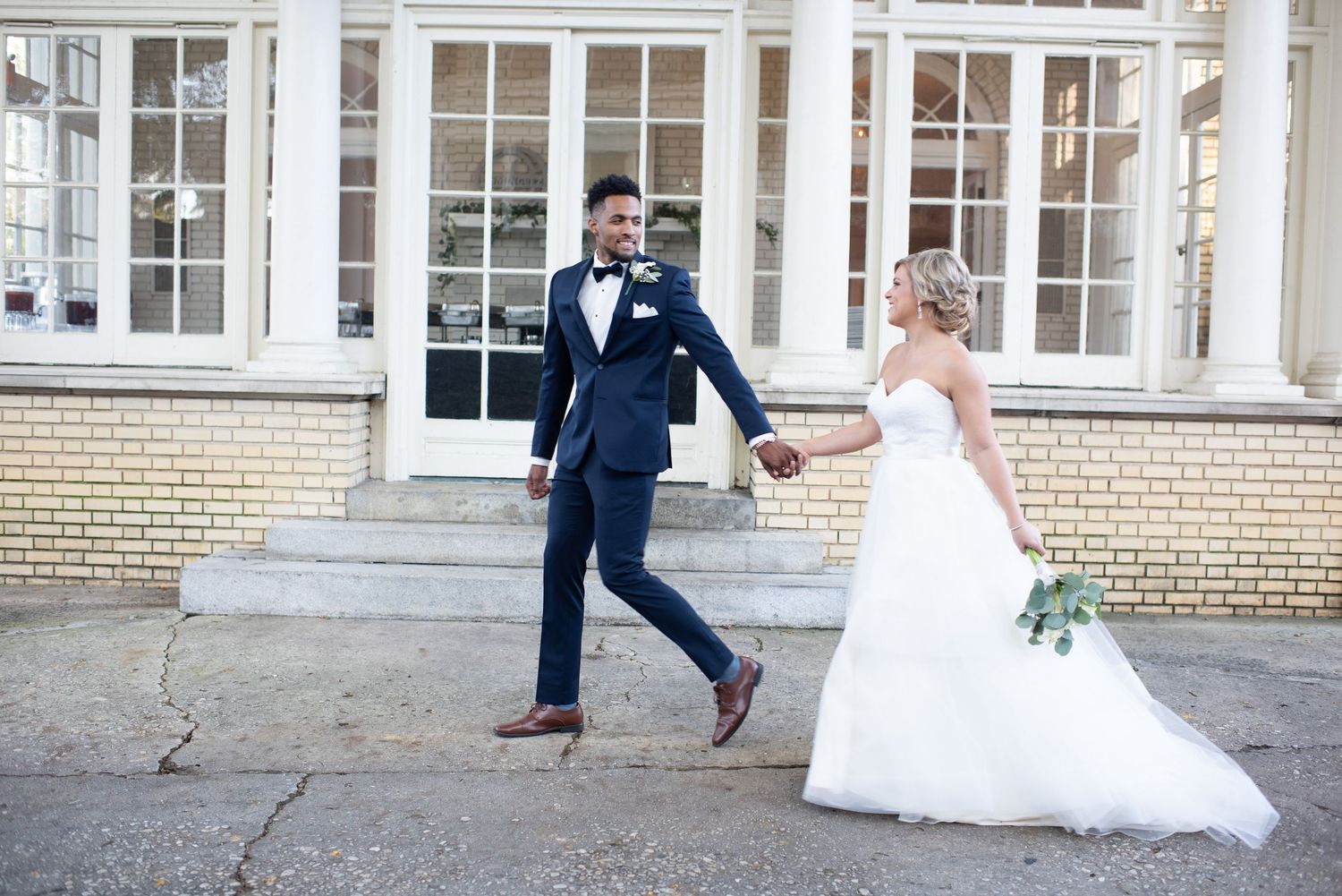 Stylish groom in navy suit leads bride by the hand outside a brick building.