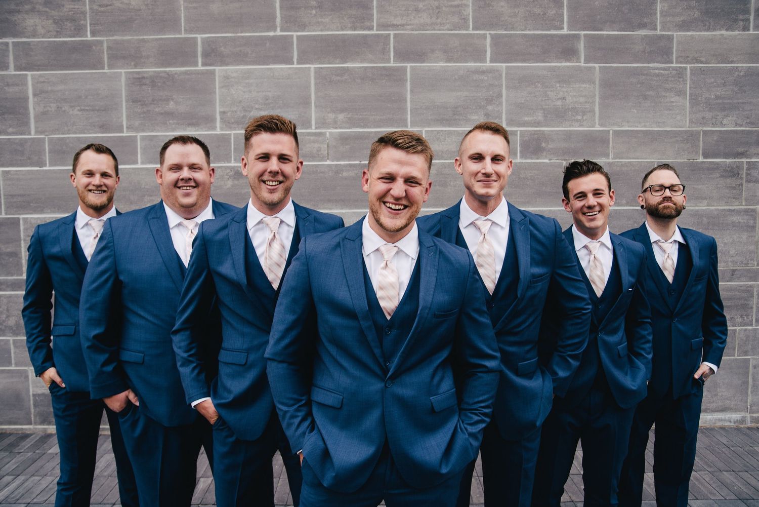 Groom and six groomsmen in matching navy blue suits with light pink ties posing in front of a gray brick wall.